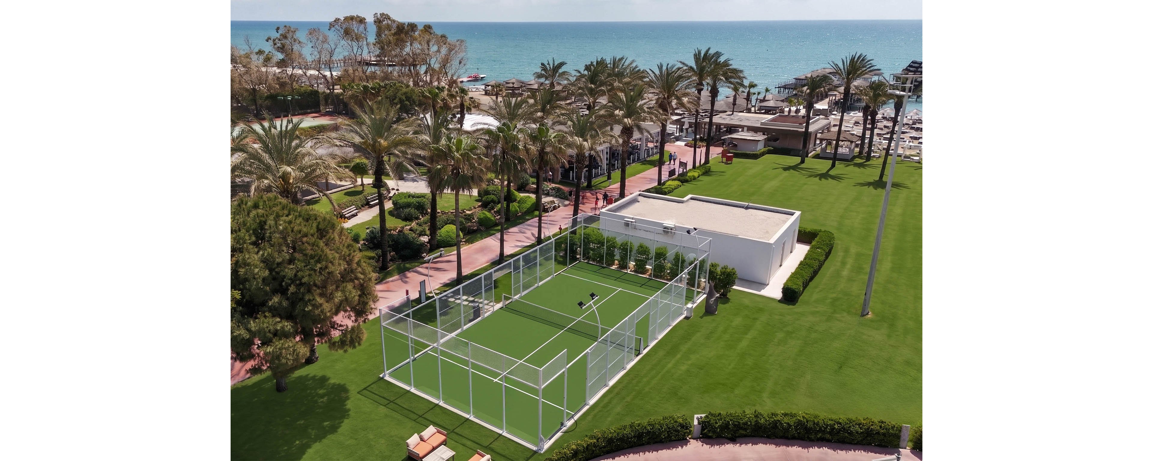 An aerial view shows a modern padel court set within a lush green lawn at a coastal resort. The fenced court sits beside a white rectangular building and landscaped pathways lined with palm trees. In the background, guests relax near the beach and swimming areas along the shoreline. The bright sunlight and clear blue sea create a vibrant, summery atmosphere.