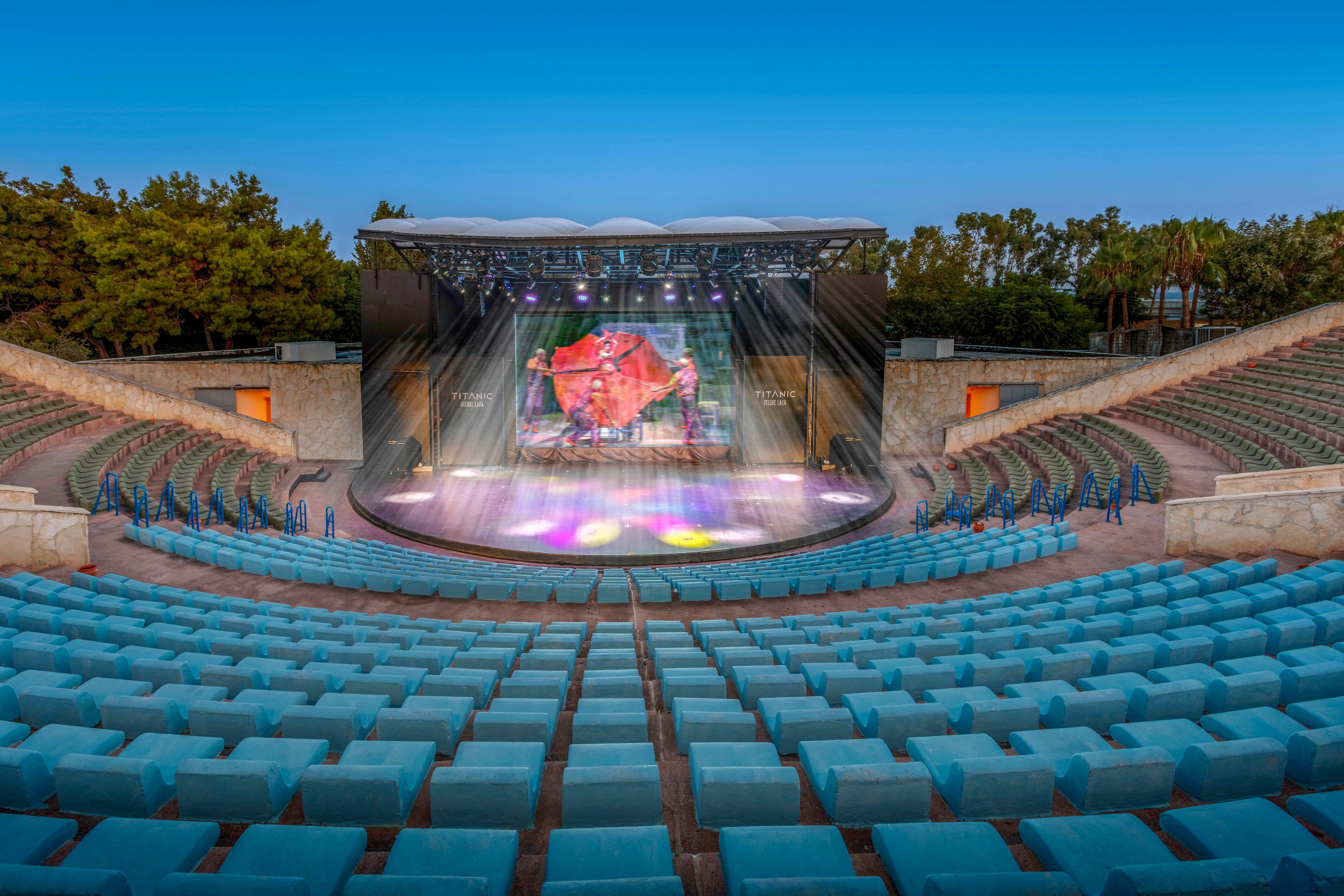 An open-air amphitheater features rows of turquoise seats facing a central stage. The stage is lit with colorful spotlights and a large screen displaying abstract imagery. Stone walls and surrounding trees frame the venue under a clear blue sky. The scene is empty of people, emphasizing the architectural layout and performance space.