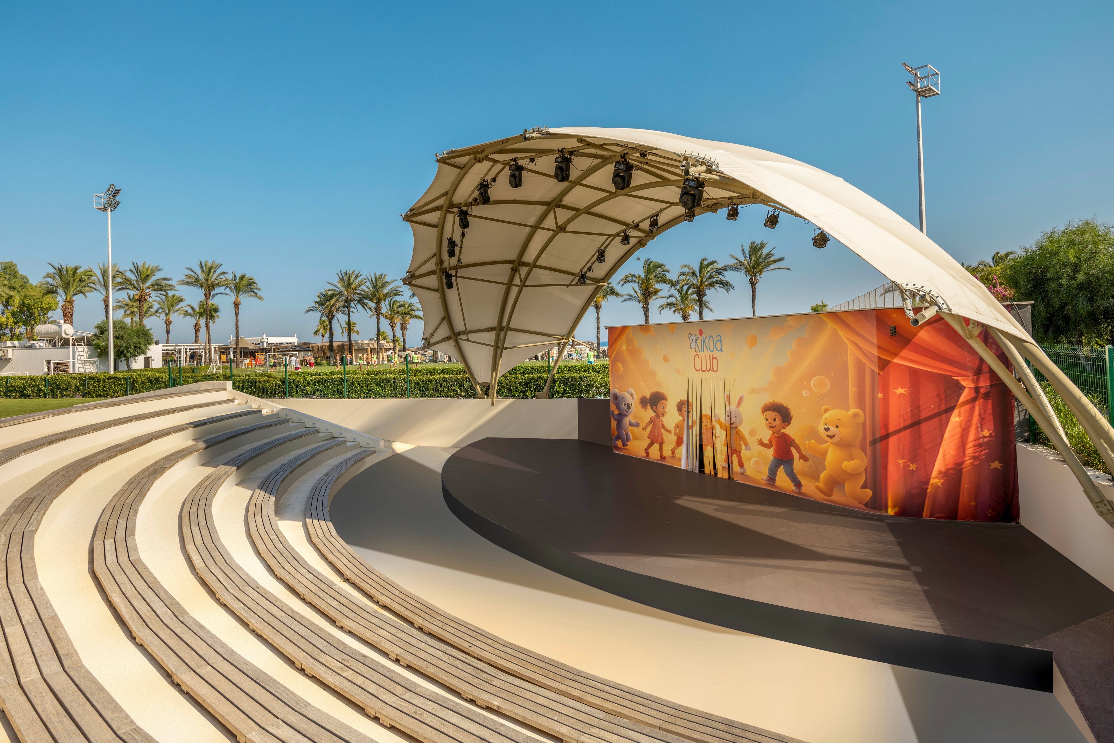 An open-air amphitheater features curved tiered seating facing a small covered stage. The stage has a modern white canopy roof with lighting fixtures suspended underneath. A colorful backdrop with cartoon-style characters decorates the rear of the stage. Palm trees, greenery, and clear blue sky create a warm, sunny resort-like atmosphere.