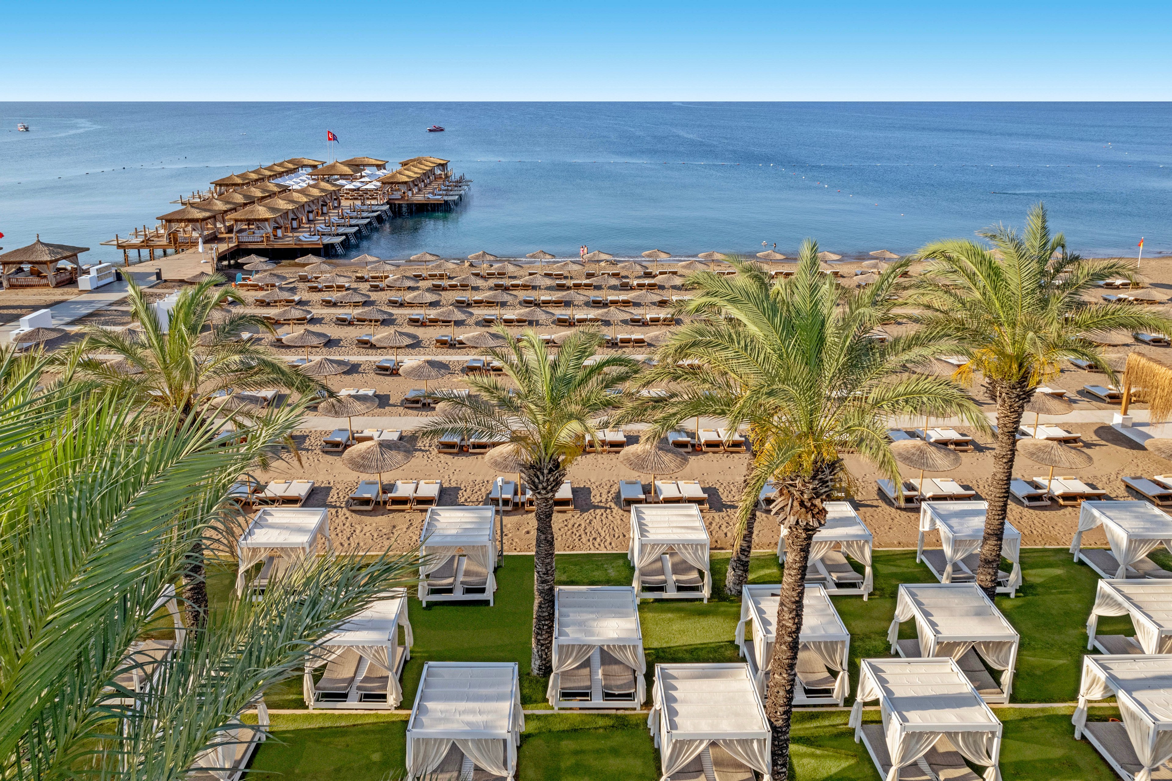 A luxury beachfront resort features rows of white cabanas and sun loungers arranged neatly along the sandy shore. Tall palm trees line the foreground, adding a tropical feel to the scene. Wooden piers extend into the calm blue sea, with small structures and flags visible at the end. The clear sky and tranquil water create a serene vacation atmosphere focused on relaxation and seaside leisure.