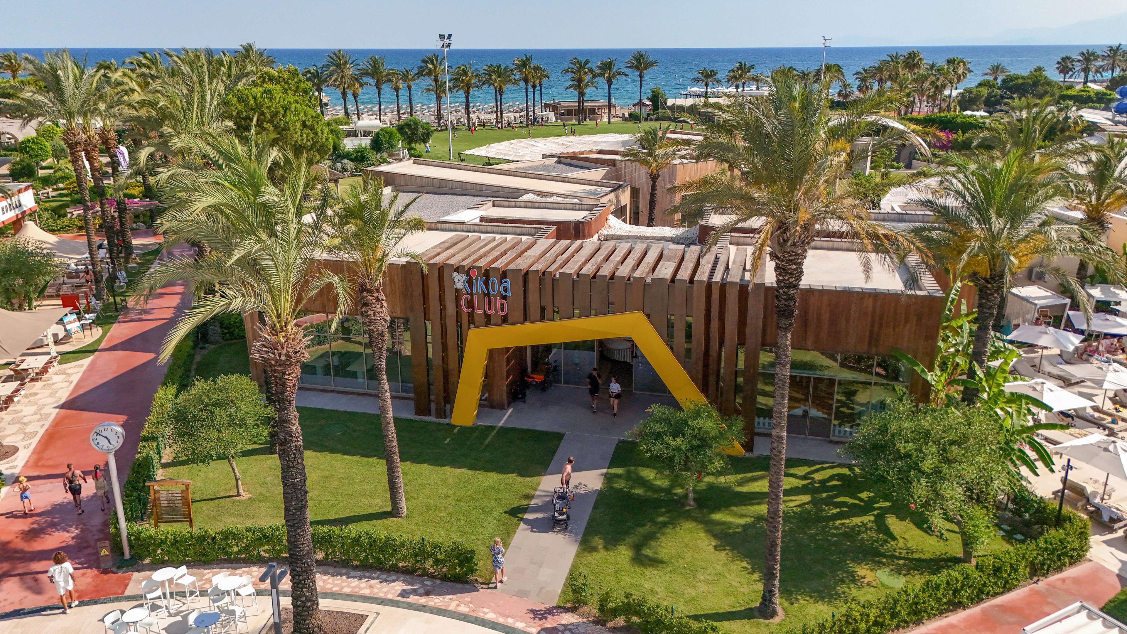 A modern kids club building sits at the center of a lush seaside resort. The structure features wooden paneling and a bold yellow entrance frame, surrounded by green lawns and tall palm trees. People walk along red and white pathways, with sun loungers and umbrellas visible nearby. In the background, the blue sea and clear sky emphasize a sunny, relaxed vacation atmosphere.