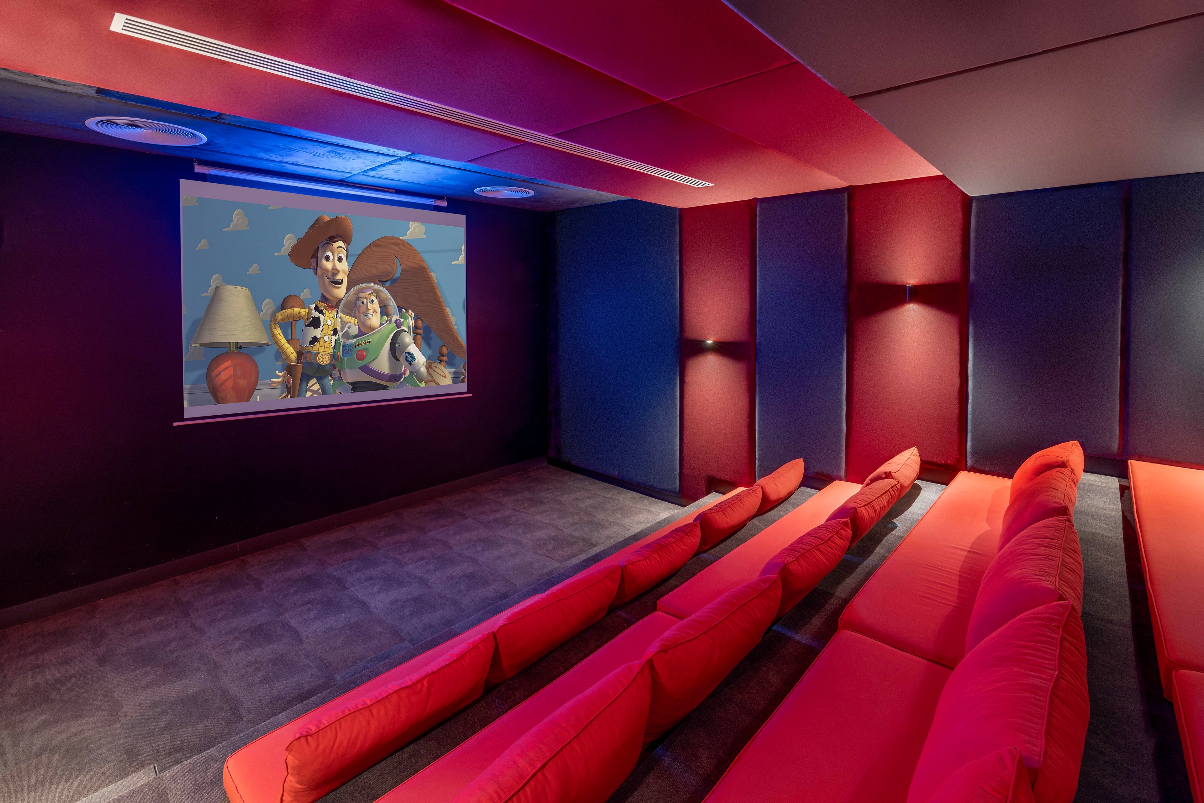 A small private home theater features rows of bright red cushioned seats facing a projection screen. An animated movie with cartoon characters is playing on the screen at the front of the room. The walls are covered with dark blue and red acoustic panels, creating a cozy, immersive environment. Recessed ceiling lights and subtle blue accent lighting enhance the cinematic atmosphere.