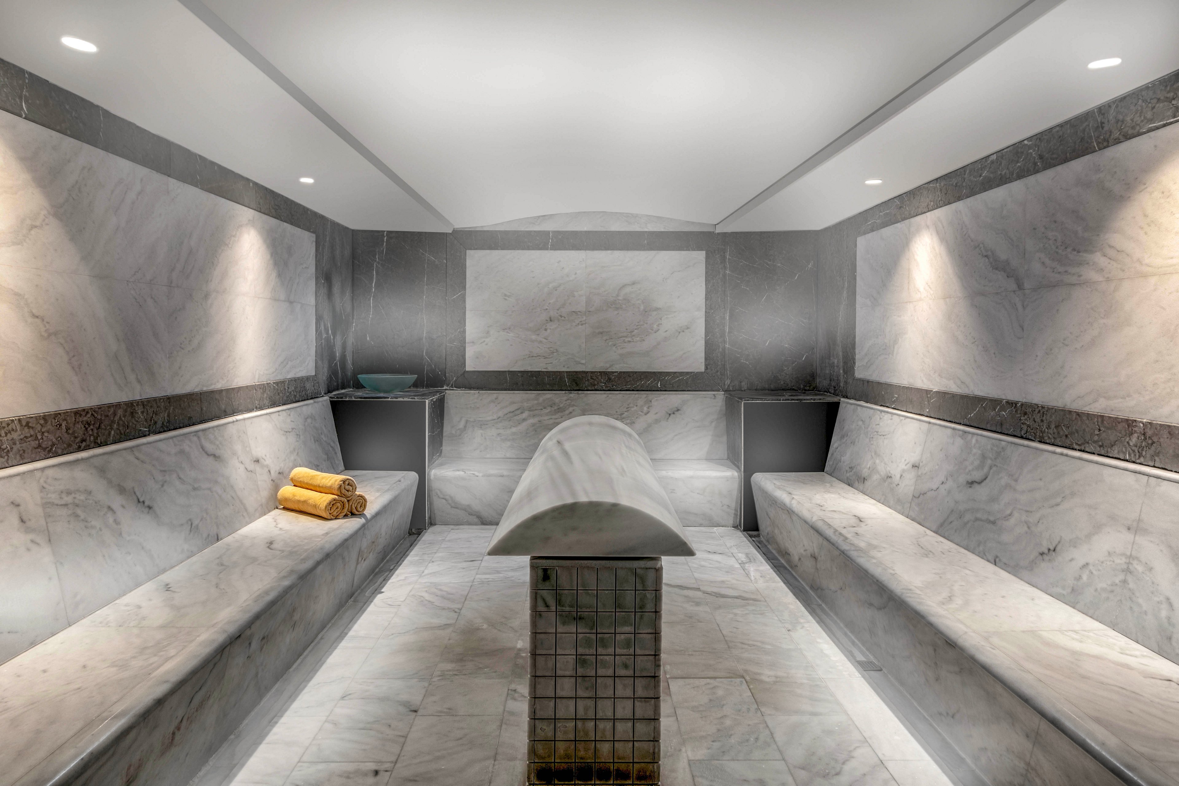 Elegant marble steam room with built-in benches and a central stone table creates a serene spa environment. Soft recessed ceiling lights illuminate the smooth gray and white stone surfaces. Rolled yellow towels add a subtle accent of color on one bench. The space appears clean, modern, and designed for relaxation and wellness.