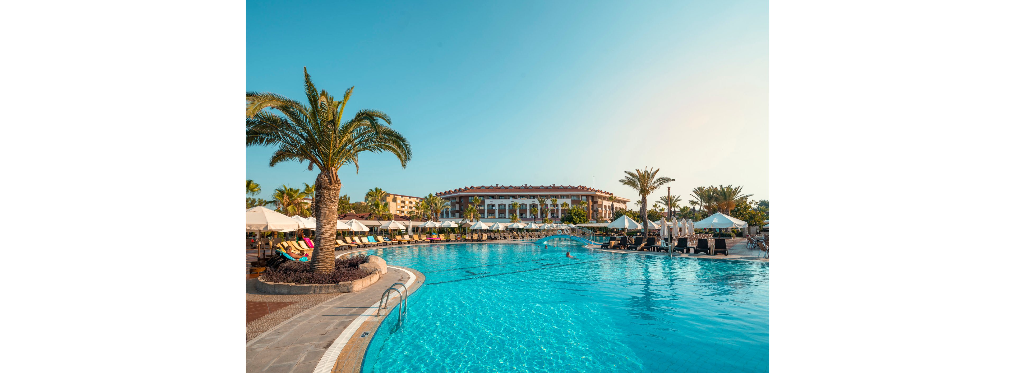 A large, clear blue swimming pool stretches across the foreground of a luxury resort. Sun loungers with colorful cushions and umbrellas line the poolside under tall palm trees. A grand hotel building with multiple arches and balconies stands in the background. The scene is bathed in warm sunlight, suggesting a relaxing vacation atmosphere in a tropical or coastal destination.