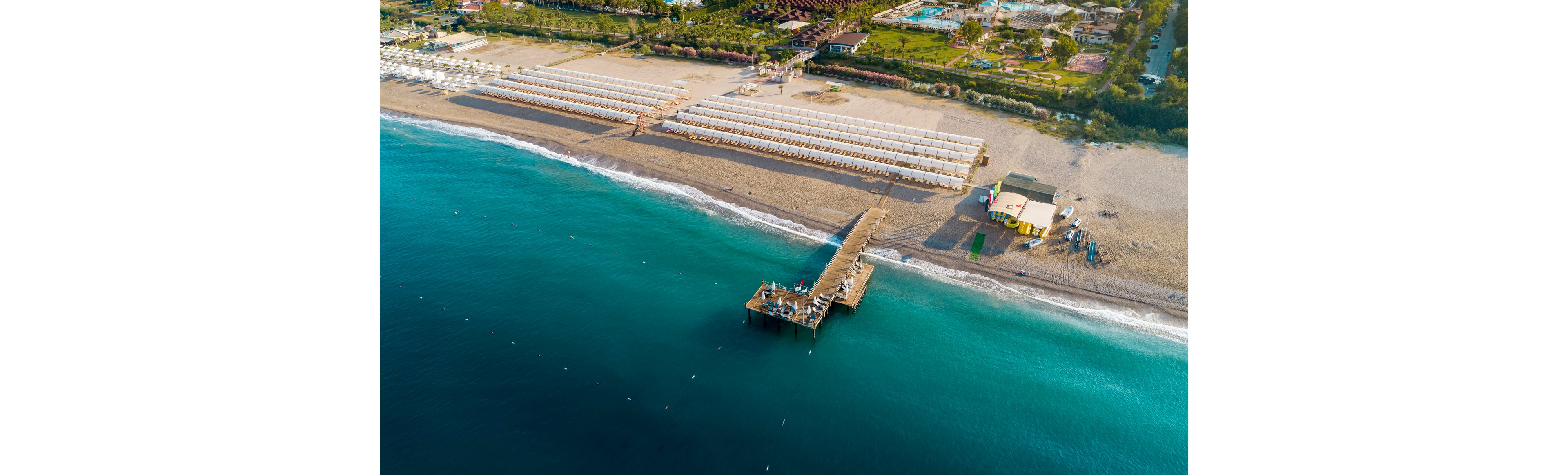 A long wooden pier extends into clear turquoise water from a sandy beach. Rows of neatly arranged sun loungers and umbrellas line the shore, indicating a well-organized beach resort. A few small buildings and landscaped areas are visible behind the beach, suggesting additional facilities and accommodations. The scene is calm and orderly, with gentle waves meeting the coastline under bright daylight.