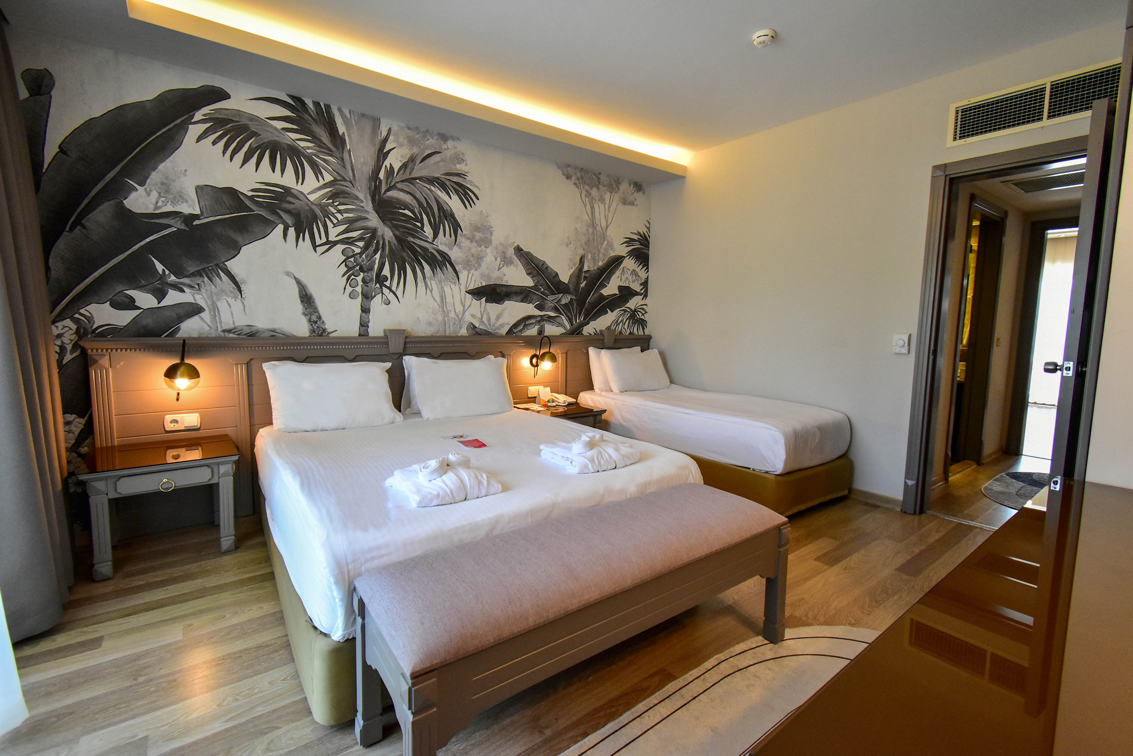 A contemporary hotel room features a large double bed and a single bed neatly made with white linens. The space has wooden flooring, warm lighting, and a monochrome tropical wall mural behind the beds. Two bedside tables with lamps flank the main bed, and folded bathrobes are placed on top. An open doorway leads to an adjacent area, enhancing the sense of depth and comfort in the room.