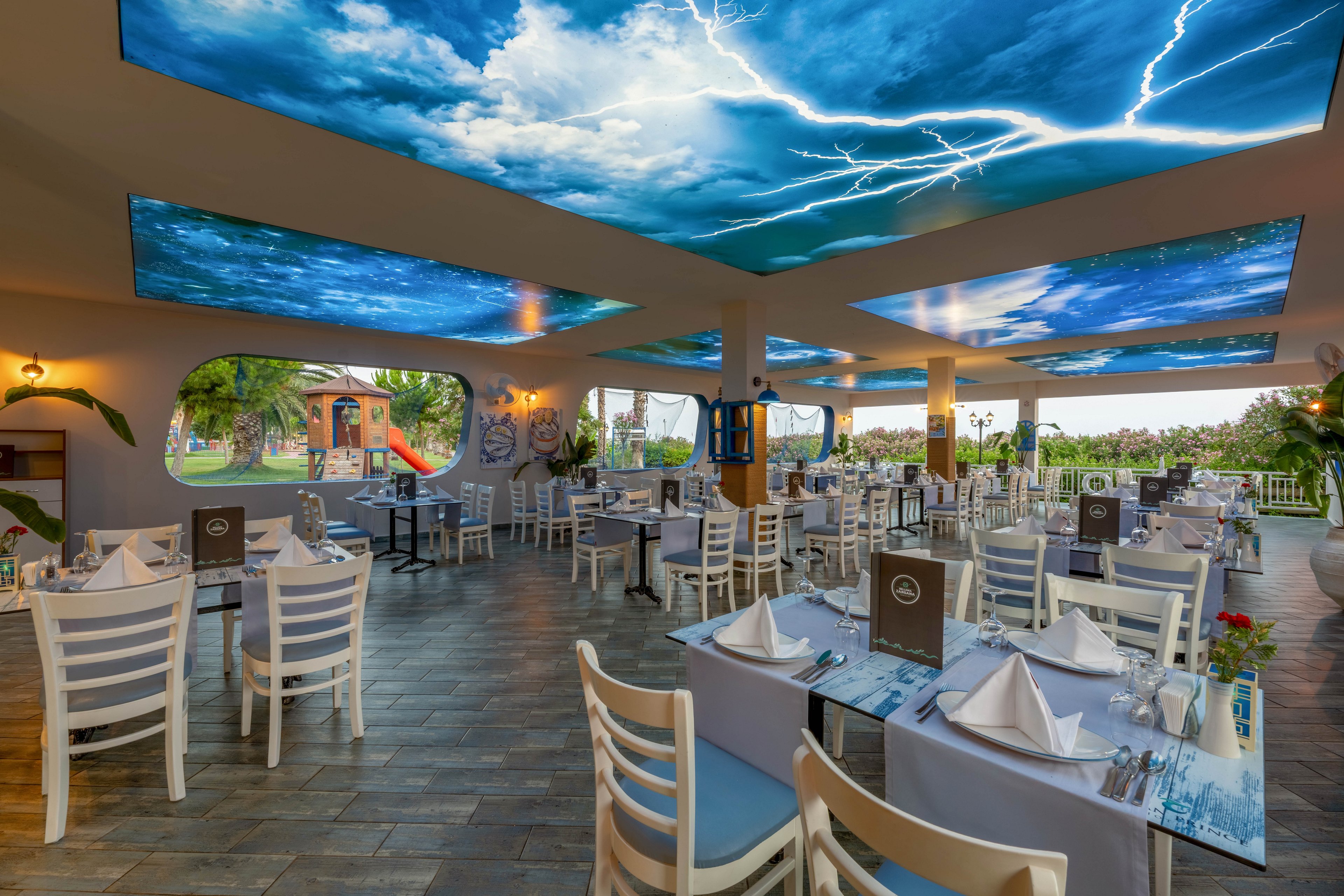 A spacious open-air restaurant features neatly arranged white tables and chairs. The ceiling panels display vivid images of a stormy sky with clouds and lightning, creating a dramatic visual effect. Large windows and openings reveal greenery and an outdoor playground in the background. The setting appears prepared for guests with folded napkins, menus, and table decor in place.