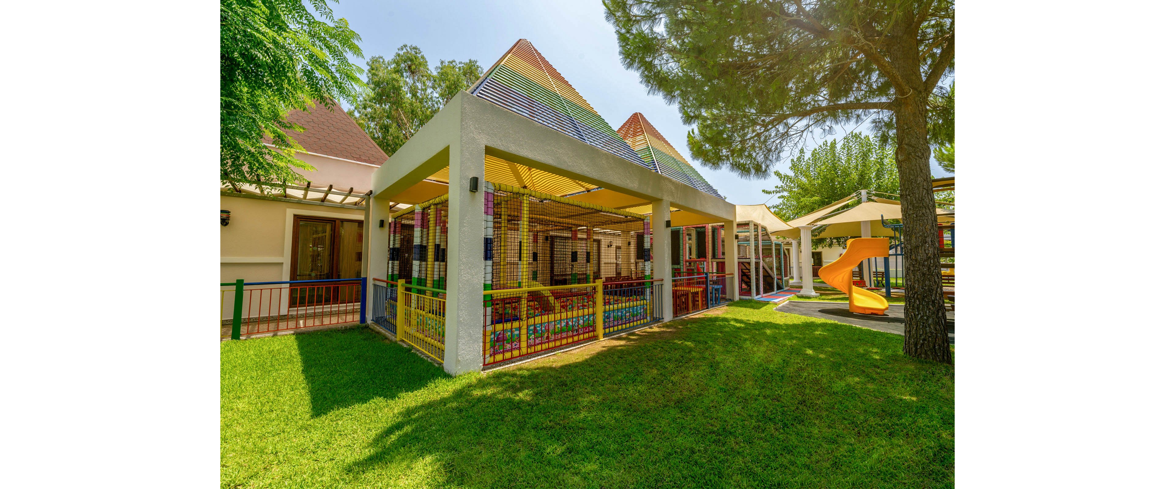 A colorful outdoor playground structure stands in the yard of a daycare or preschool. The play area is covered by a geometric roof with bright stripes and surrounded by safety fencing. A slide and additional play equipment are visible in the background under the shade of trees. Lush green grass and clear sunlight create a cheerful, inviting atmosphere for children.