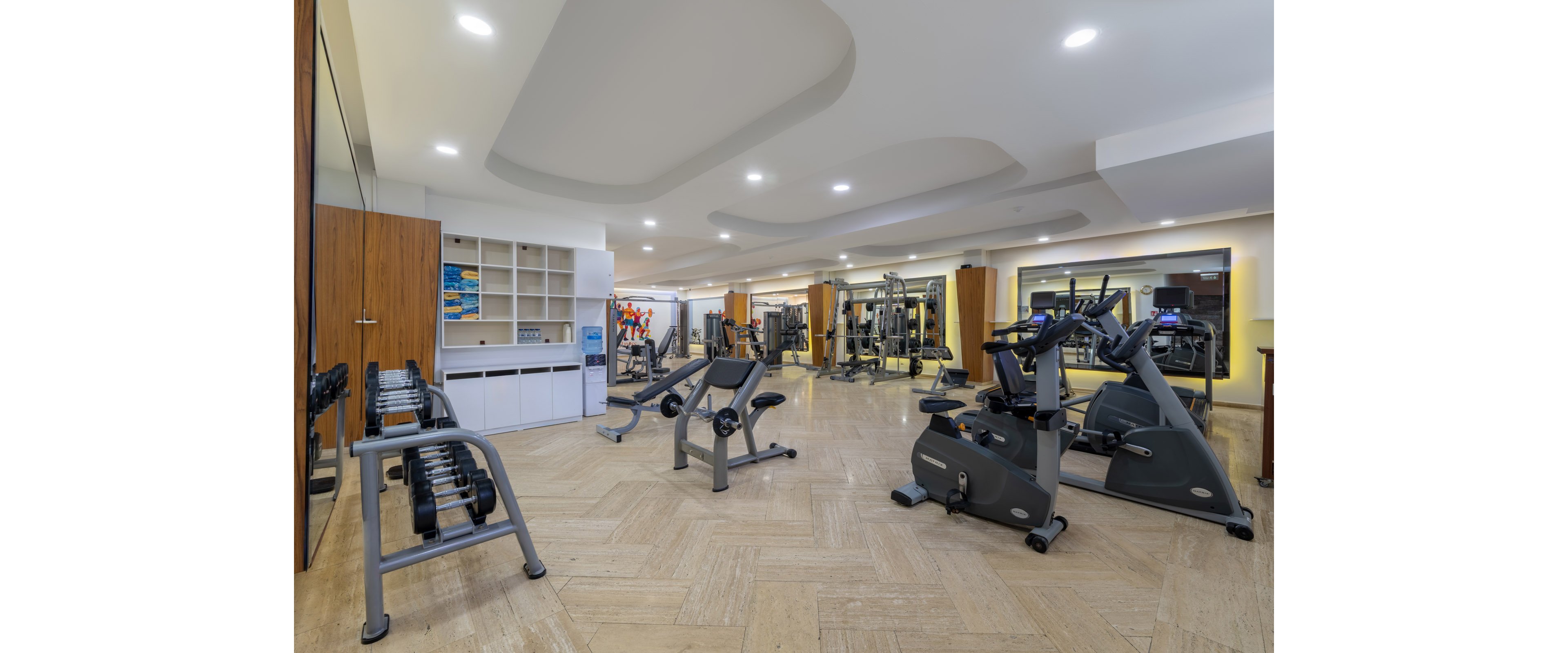 A modern fitness center features a variety of exercise equipment neatly arranged in a bright, spacious room. Stationary bikes and elliptical machines are positioned in the foreground, with weight benches and dumbbells to the side. Additional strength-training machines and mirrors line the back wall, enhancing the sense of depth and space. Recessed ceiling lights and light-colored flooring create a clean, contemporary atmosphere suitable for workouts.