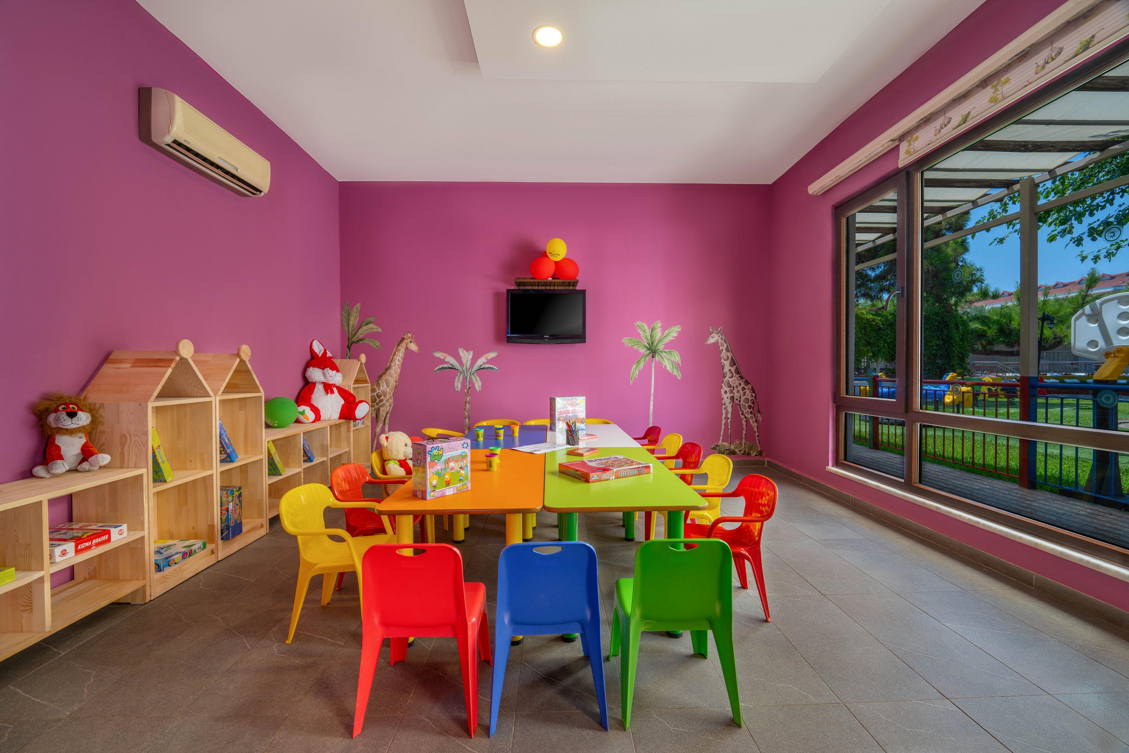 Brightly colored children's playroom with a central table surrounded by small plastic chairs in red, yellow, blue, and green. The room features a purple wall, wooden shelves filled with toys and books, and playful animal decorations. Large windows on one side look out onto an outdoor playground area with greenery. A wall-mounted television and ceiling lighting complete the modern, cheerful space designed for kids' activities.