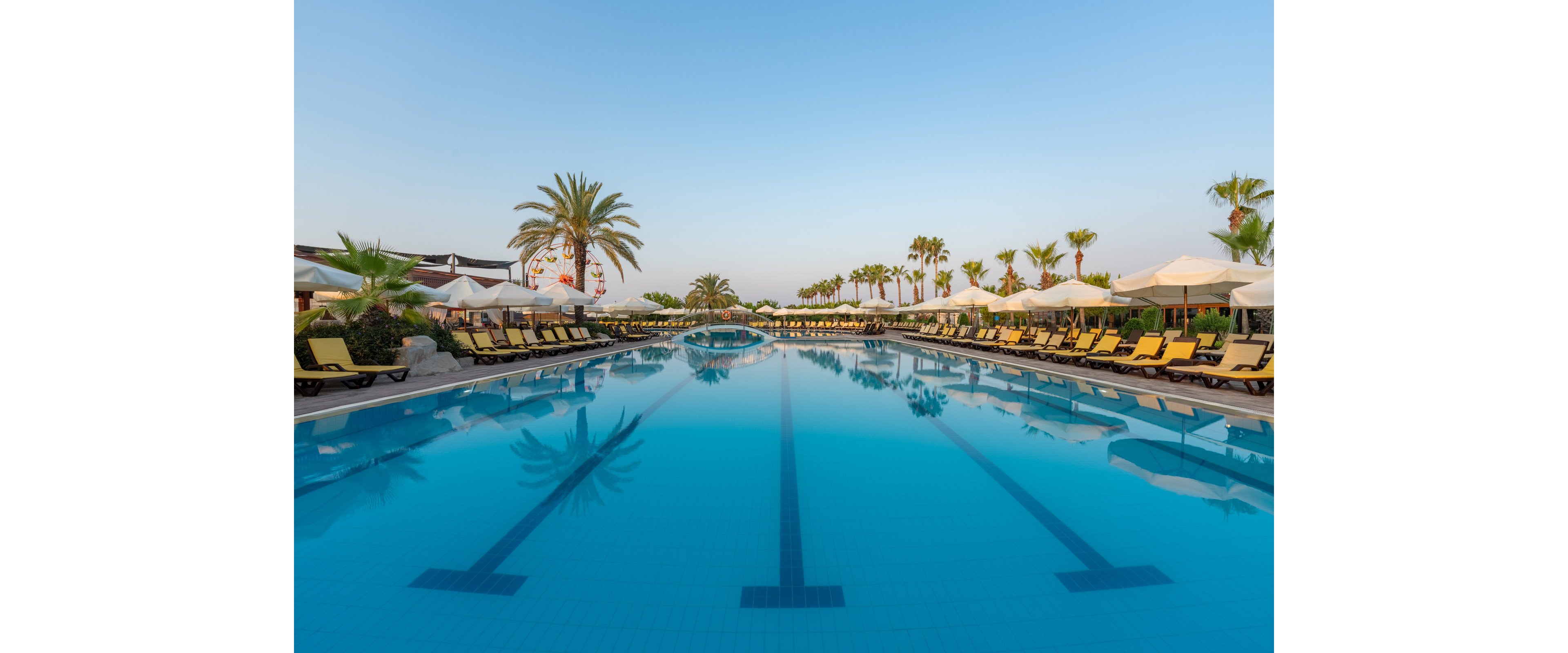 A large resort swimming pool stretches into the distance under a clear blue sky. Yellow sun loungers and white umbrellas line both sides of the pool, creating a symmetrical composition. Tall palm trees frame the scene, reflecting in the calm water. The tranquil atmosphere suggests an early morning or late afternoon setting at a vacation destination.