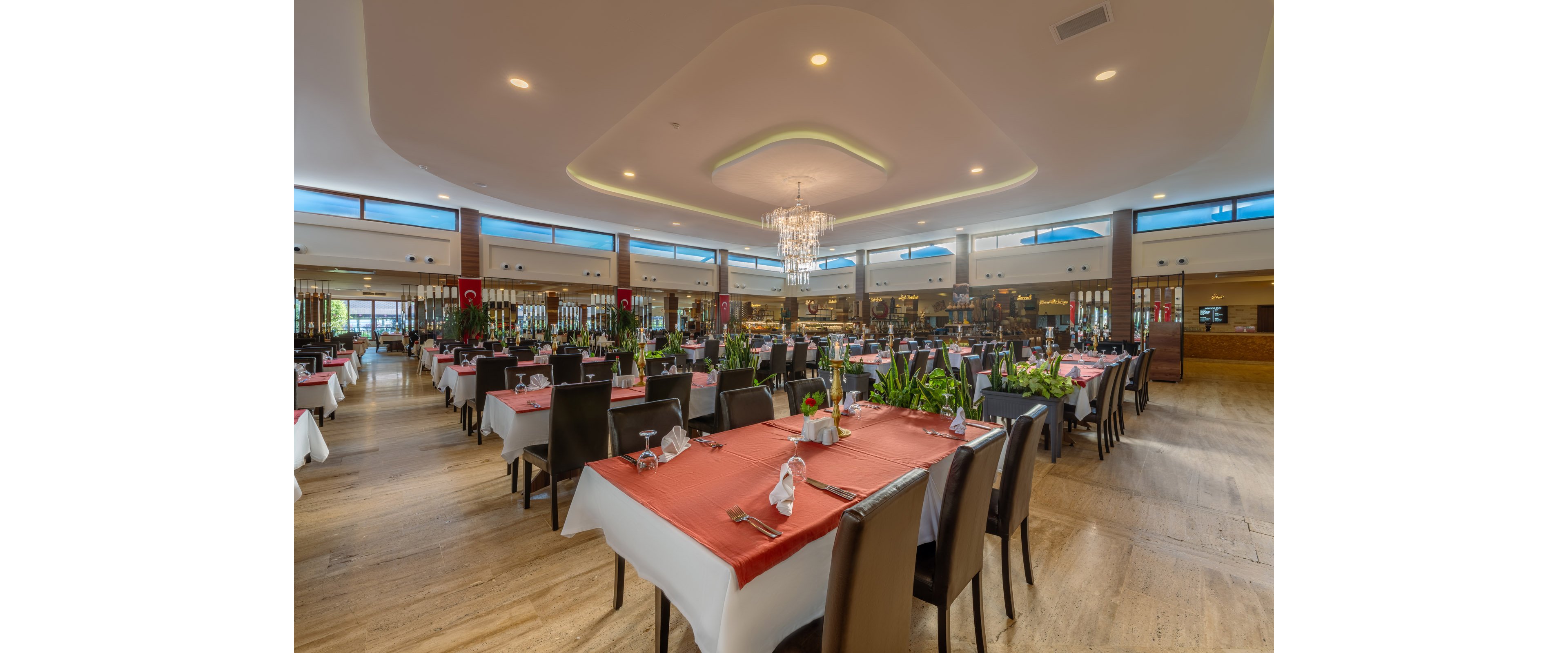A spacious, upscale restaurant dining room is shown with rows of neatly arranged tables covered in white cloths and rust-colored runners. Dark wooden chairs surround each table, which are set with glassware, folded napkins, and cutlery. Green plants and decorative centerpieces add freshness between the seating areas. Large windows and a central chandelier provide bright, inviting lighting in the modern interior.