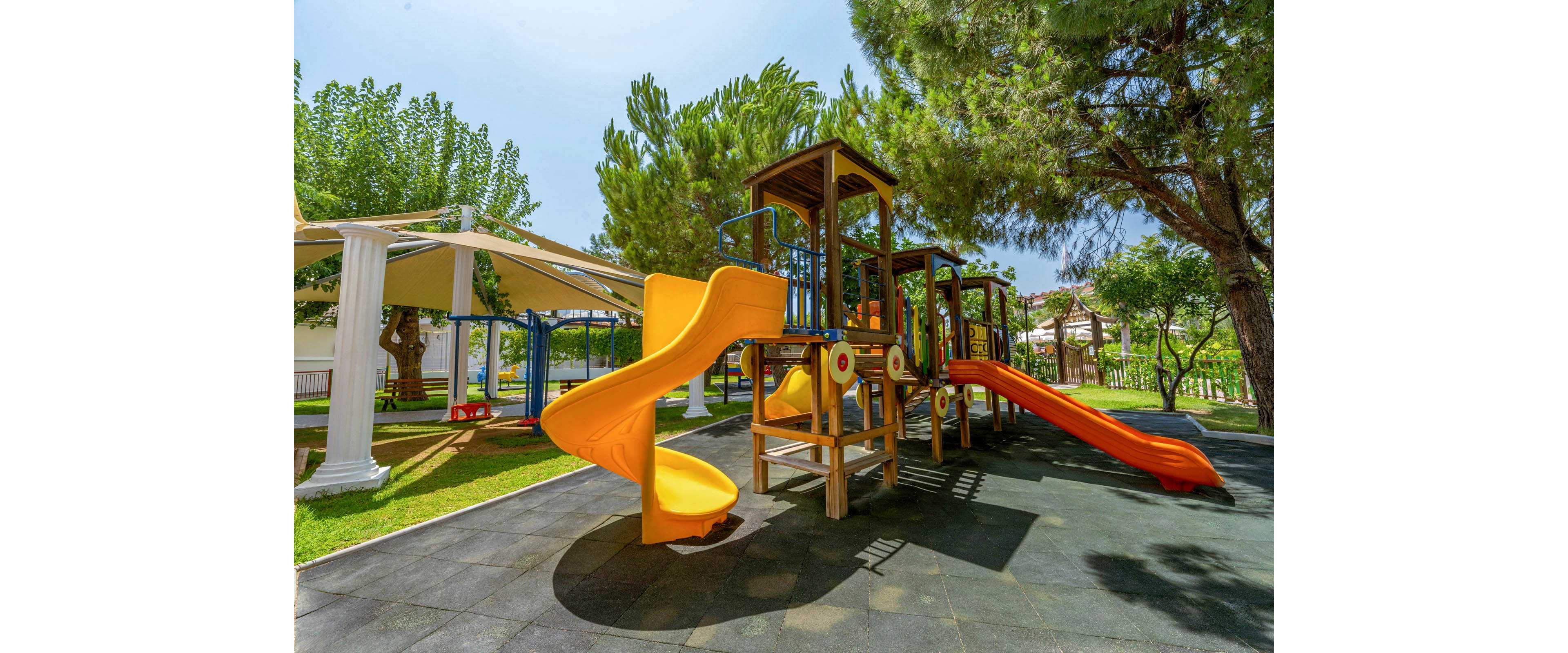 A bright outdoor playground features multiple slides and climbing structures under tall green trees. The main playset includes yellow and orange tube and wave slides connected by wooden platforms and metal railings. Soft safety flooring surrounds the equipment, and a shaded seating or play area is visible in the background. The scene is lit by clear midday sunlight, creating a cheerful and inviting atmosphere for children.