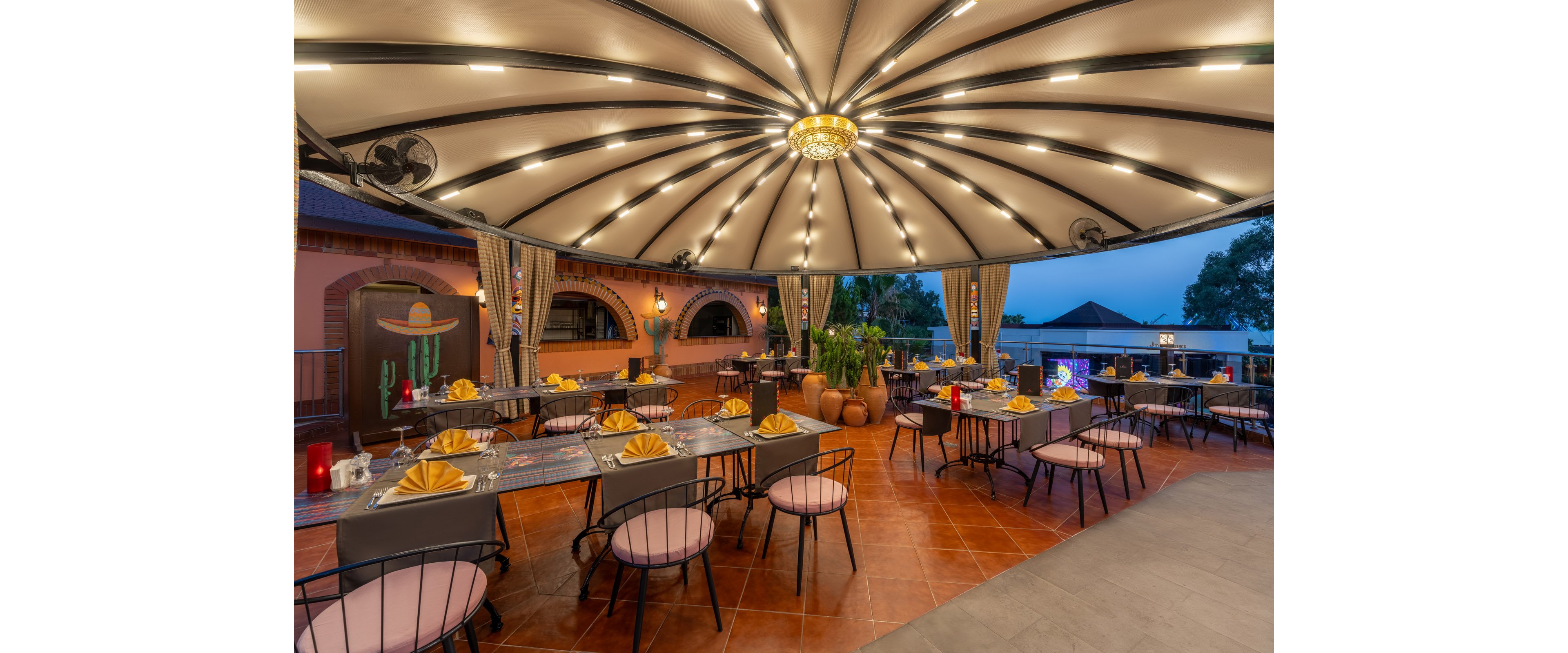 An open-air restaurant dining area is arranged with neatly set tables under a large circular canopy of warm lights. Modern metal chairs surround tables decorated with yellow folded napkins and glassware. Potted plants and draped curtains add a touch of greenery and softness to the tiled terrace floor. The scene is captured at dusk, creating a cozy, inviting atmosphere with views of surrounding rooftops and trees.