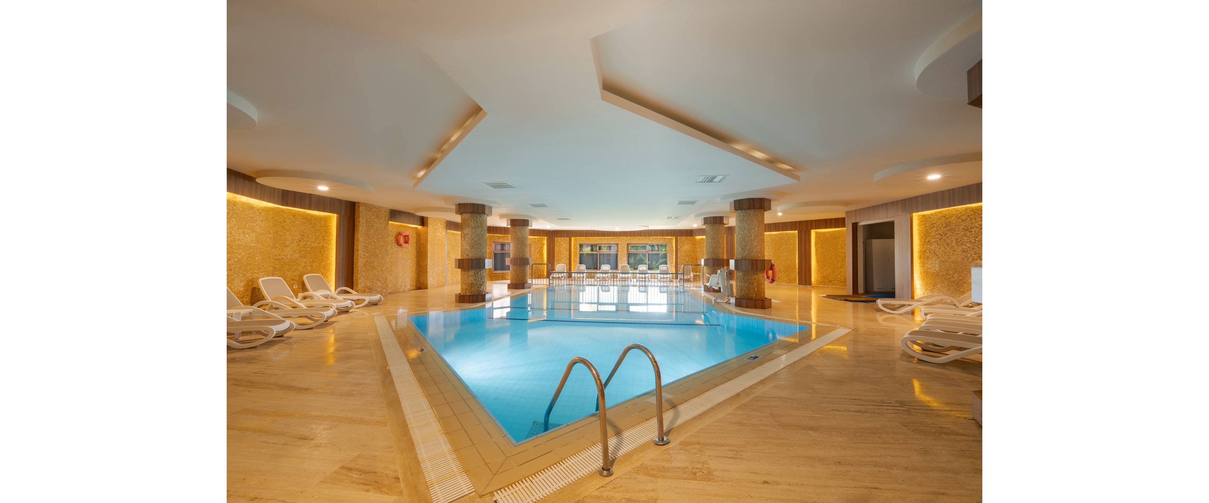 Spacious indoor swimming pool in a modern hotel or spa. The pool is surrounded by beige marble flooring, warm yellow lighting, and decorative wall panels. White lounge chairs line the sides, offering a relaxing place to rest between swims. Large windows at the far end let in natural light, enhancing the calm and inviting atmosphere.