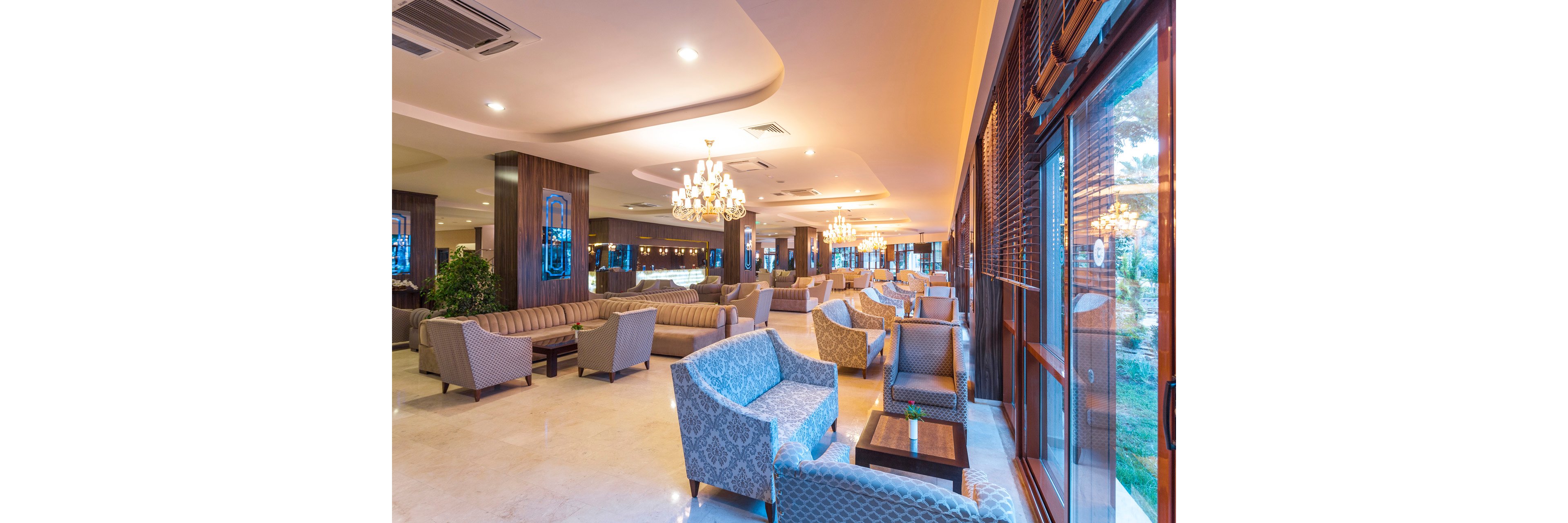 Spacious hotel lobby lounge featuring multiple seating areas with patterned armchairs and long sofas. Warm lighting from ornate chandeliers creates a welcoming and upscale atmosphere. Large windows along one side let in natural light and offer views of greenery outside. Polished floors and wooden wall accents contribute to the modern, luxurious interior design.