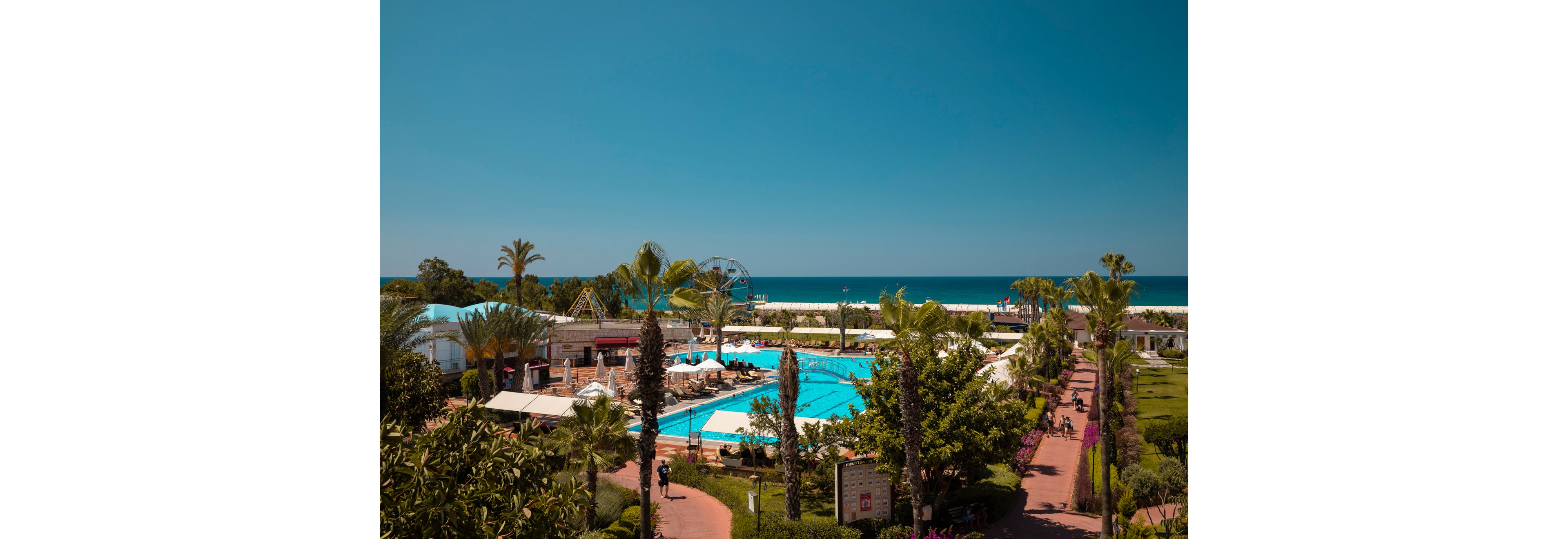 A large outdoor swimming pool sits at the center of a seaside resort. Palm trees, landscaped gardens, and walking paths surround the pool area, creating a lush vacation atmosphere. In the background, amusement rides and cabanas are visible near the shoreline. The calm blue ocean and clear sky complete the tranquil holiday setting.