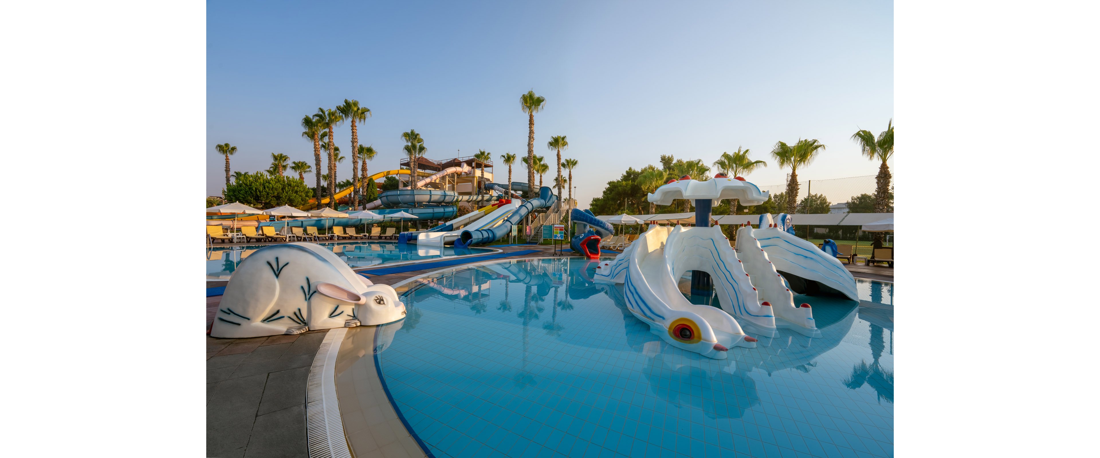 An outdoor water park features shallow pools with playful animal-shaped structures and slides. Large curved water slides and stair towers rise in the background, surrounded by palm trees and sun loungers. The calm blue water reflects the bright structures in the early morning or late afternoon light. The scene suggests a family-friendly resort area designed for children’s aquatic play.