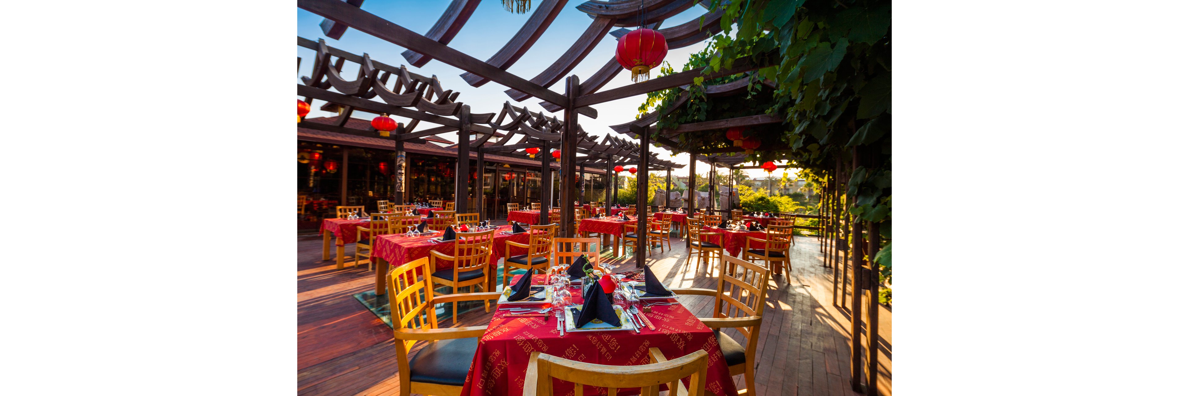 An open-air restaurant terrace is set with red tablecloths and neatly arranged place settings. Wooden chairs surround the tables under a decorative pergola structure adorned with hanging red lanterns. Sunlight creates a warm, inviting atmosphere, highlighting the greenery and scenic view in the background. The scene suggests a festive or special dining experience at sunset or late afternoon.