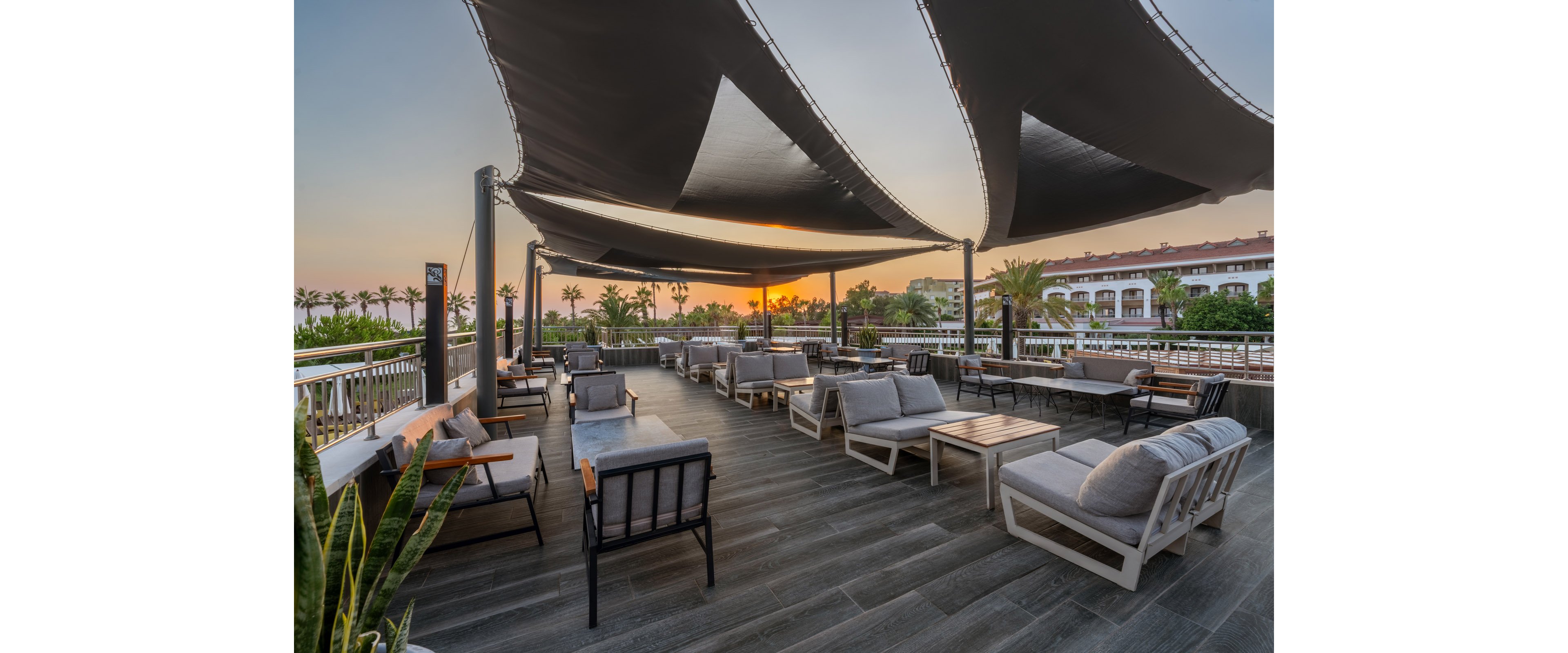 A spacious outdoor lounge terrace features contemporary sofas and low tables arranged in neat rows. Large triangular shade sails stretch overhead, framing a warm sunset on the horizon. Palm trees and resort buildings surround the deck, suggesting a coastal or vacation setting. The scene feels calm and inviting, ideal for evening relaxation or social gatherings.