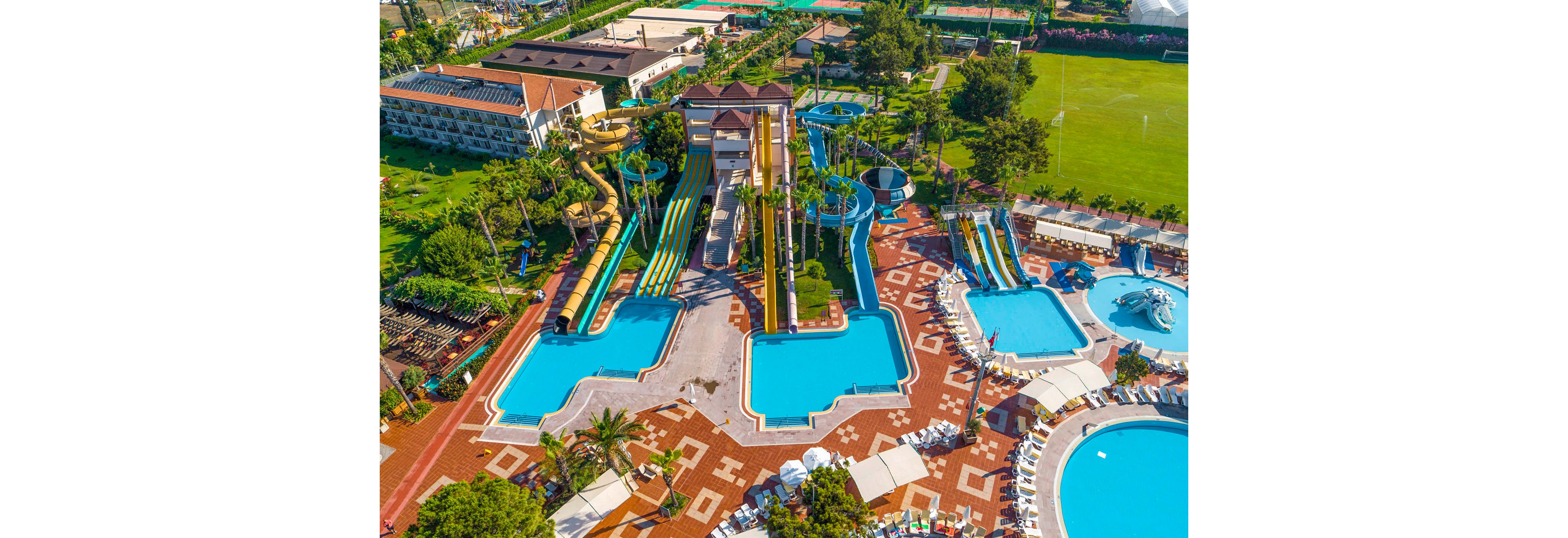 A large resort water park is captured from above, showcasing multiple pools and colorful water slides. The central structure features tall spiral and straight slides leading into rectangular blue pools. Surrounding the water attractions are sun loungers, palm trees, and tiled walkways creating a vibrant vacation atmosphere. In the background, green lawns, sports fields, and additional resort buildings are visible, emphasizing the expansive leisure facilities.