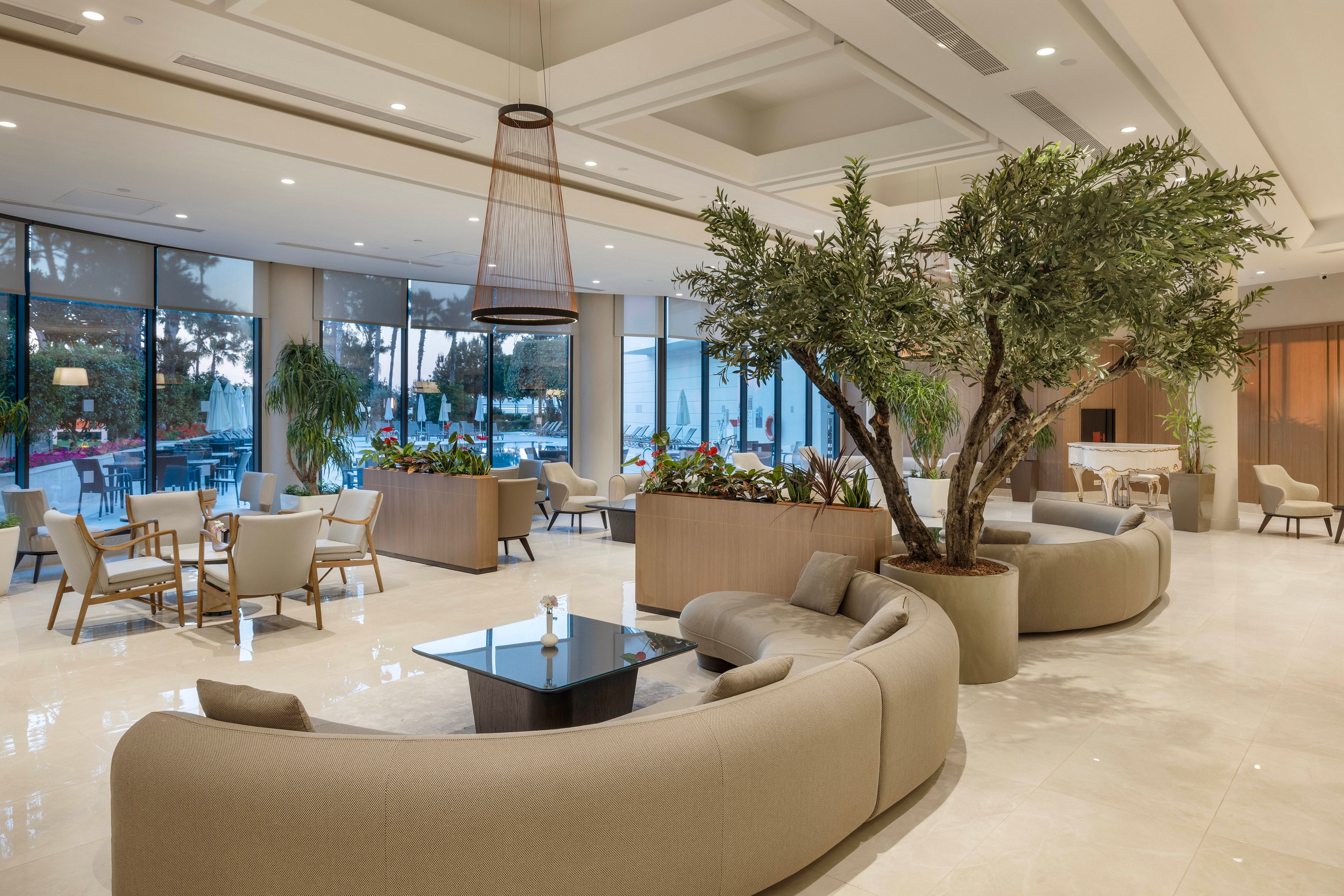 A spacious contemporary lobby features curved beige sofas arranged around a low black coffee table. Large floor-to-ceiling windows reveal an outdoor pool area and trees beyond. Neutral tones, indoor plants, and soft lighting create a calm, welcoming atmosphere. Several seating areas with armchairs and small tables provide comfortable spaces for guests to relax or socialize.