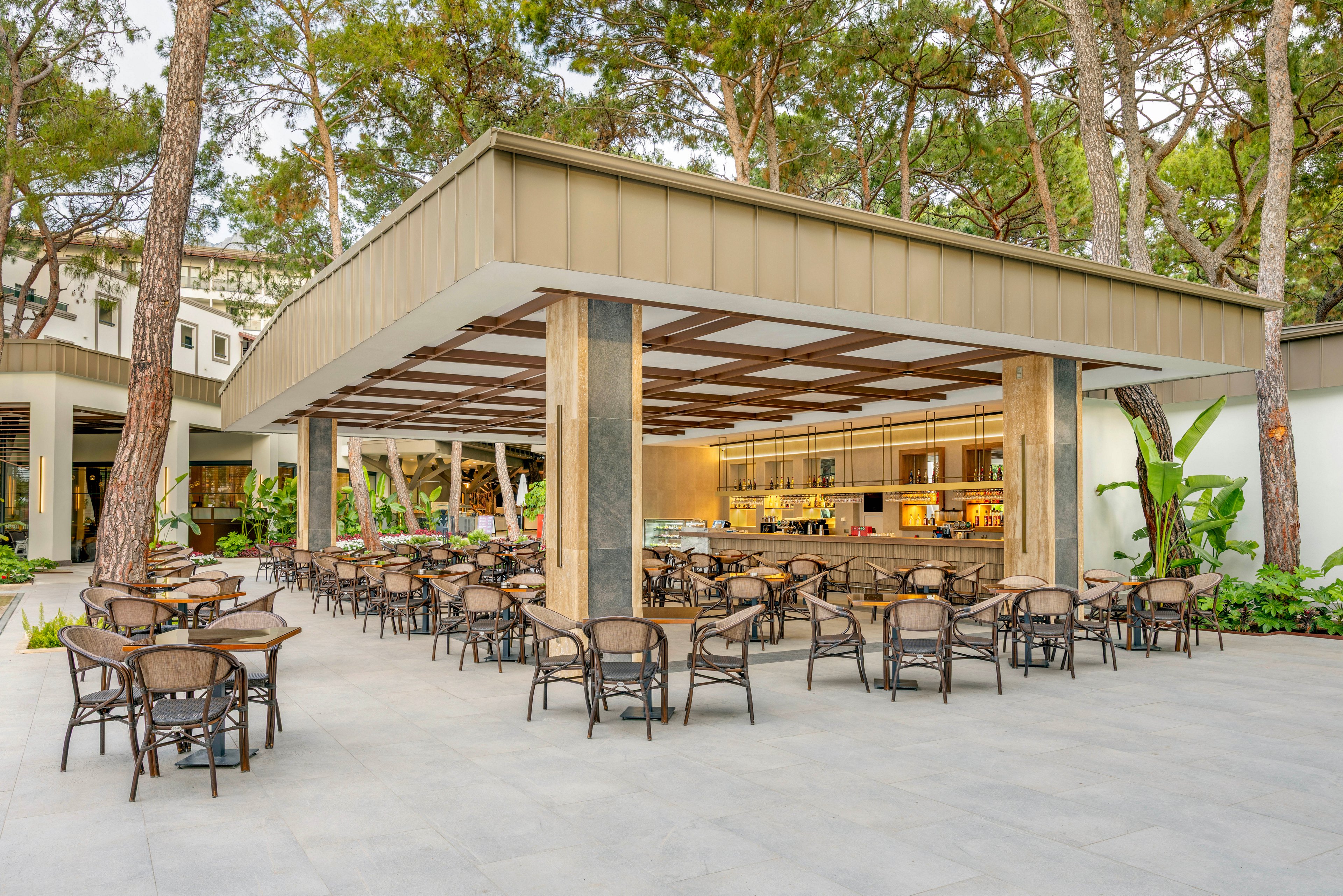 Open-air café seating area is arranged under a modern pavilion surrounded by tall trees. Numerous wicker chairs and tables are neatly organized on a wide stone terrace, facing a bar and service counter. Warm interior lighting contrasts with the natural greenery and neutral tones of the structure. The setting appears calm and inviting, ready for guests in a resort or hotel environment.