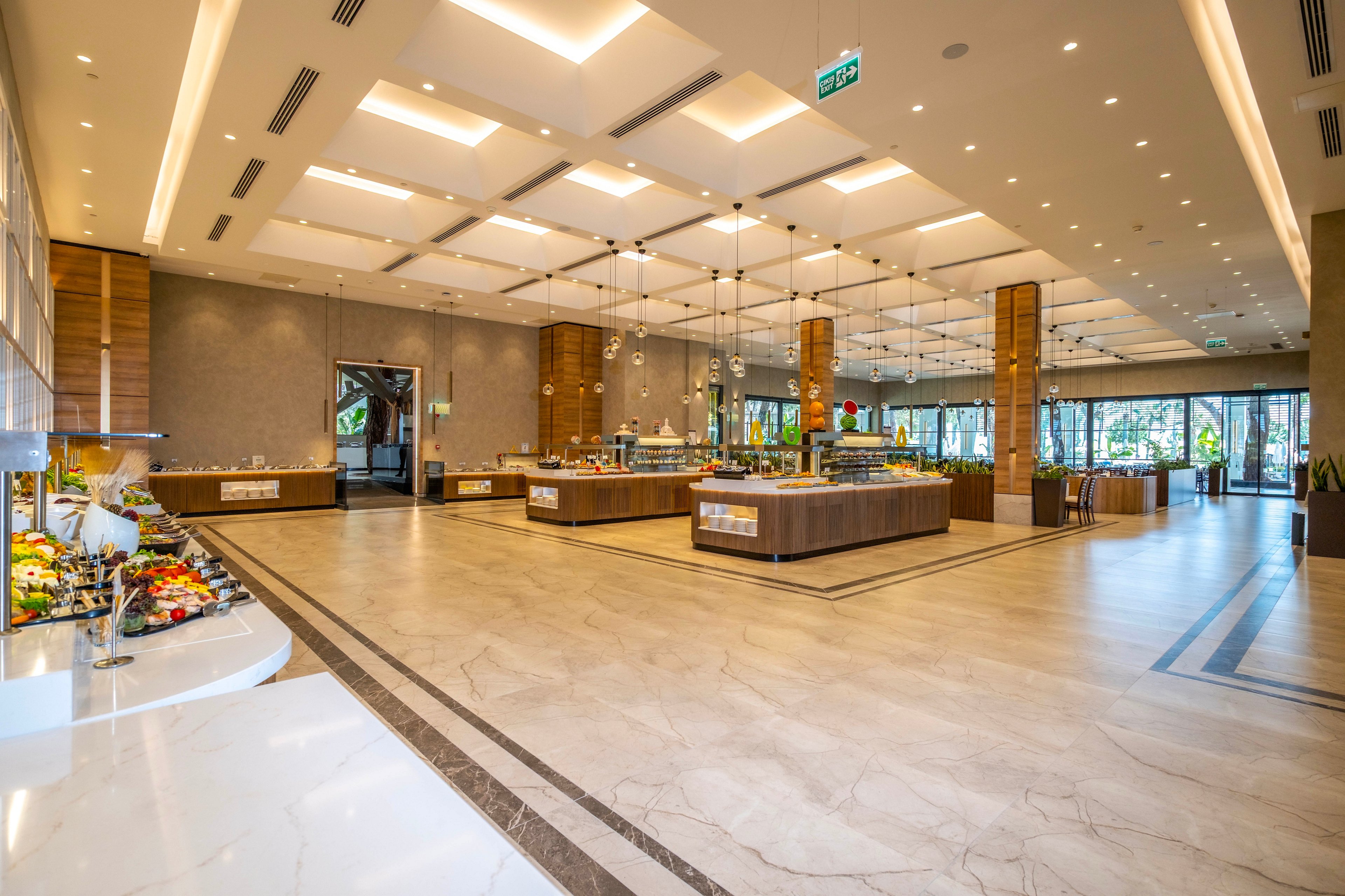 A large, modern buffet dining hall is shown with multiple food stations arranged across the room. The space features polished marble floors, warm wooden accents, and a high ceiling with recessed lighting. Various dishes and salads are displayed on counters, while large windows at the far end let in natural light and views of greenery. The overall atmosphere is clean, organized, and inviting, suggesting a high-end hotel or resort restaurant.