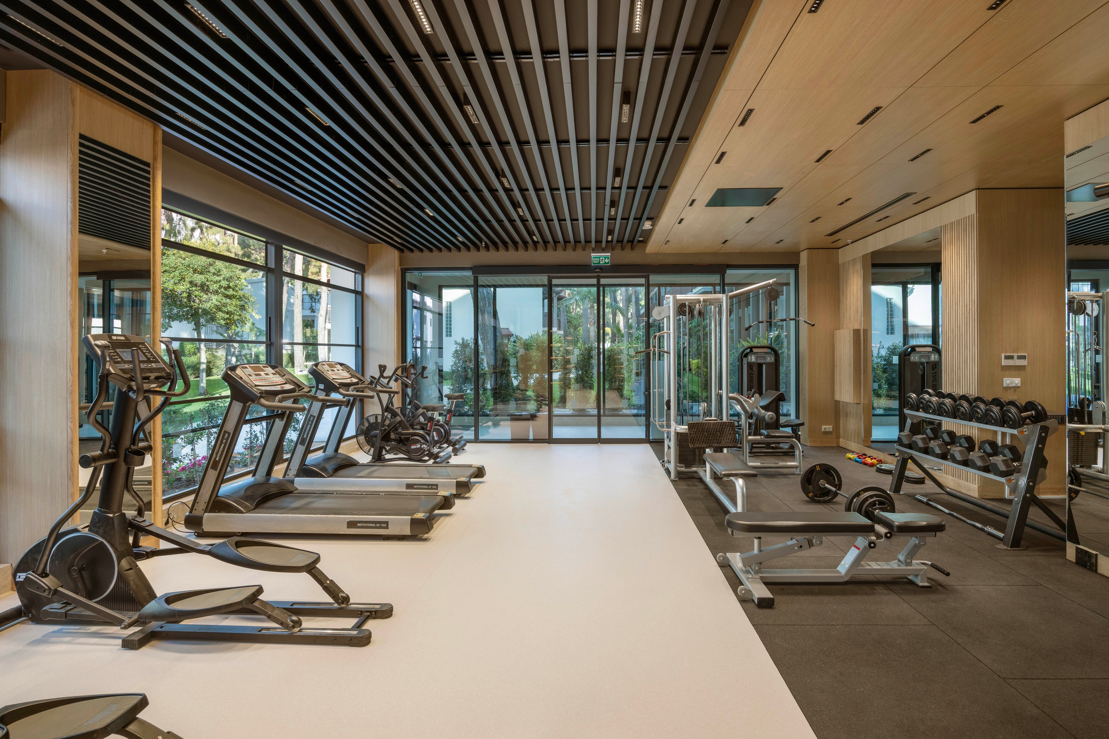 A contemporary fitness center features a variety of cardio and strength machines neatly arranged in a spacious room. Large floor-to-ceiling glass walls reveal greenery and an outdoor landscape, bringing natural light into the interior. The gym includes treadmills, ellipticals, weight benches, dumbbells, and cable machines on contrasting light and dark flooring. Warm wood paneling and a slatted ceiling create a clean, upscale atmosphere suited for exercise and wellness.