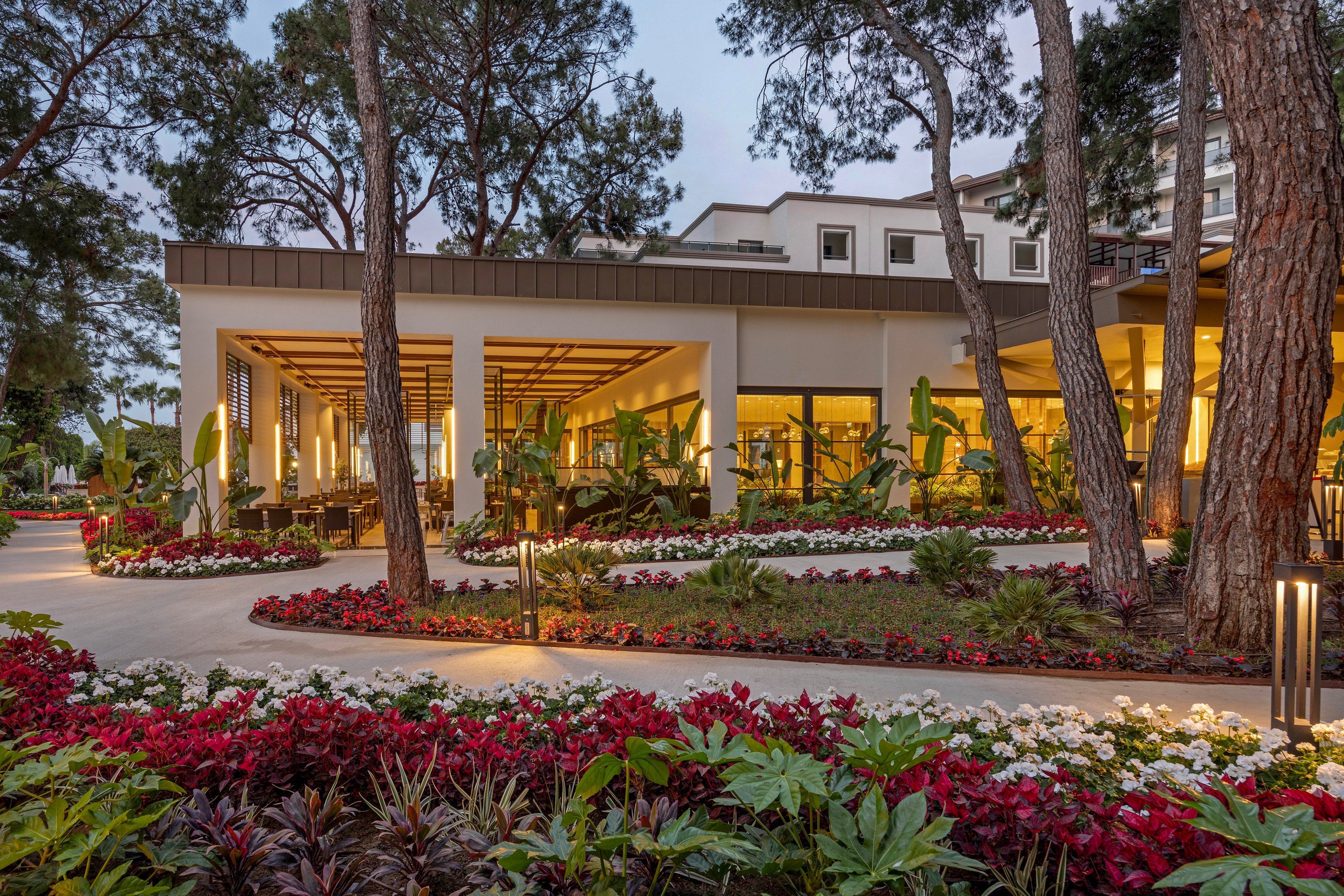 A contemporary resort-style building is surrounded by lush, manicured gardens at dusk. Warm interior lighting glows through large windows, creating an inviting atmosphere. Colorful flower beds line the curved pathways, framed by tall pine trees and tropical plants. The scene feels tranquil and upscale, suggesting a relaxing vacation or hospitality setting.