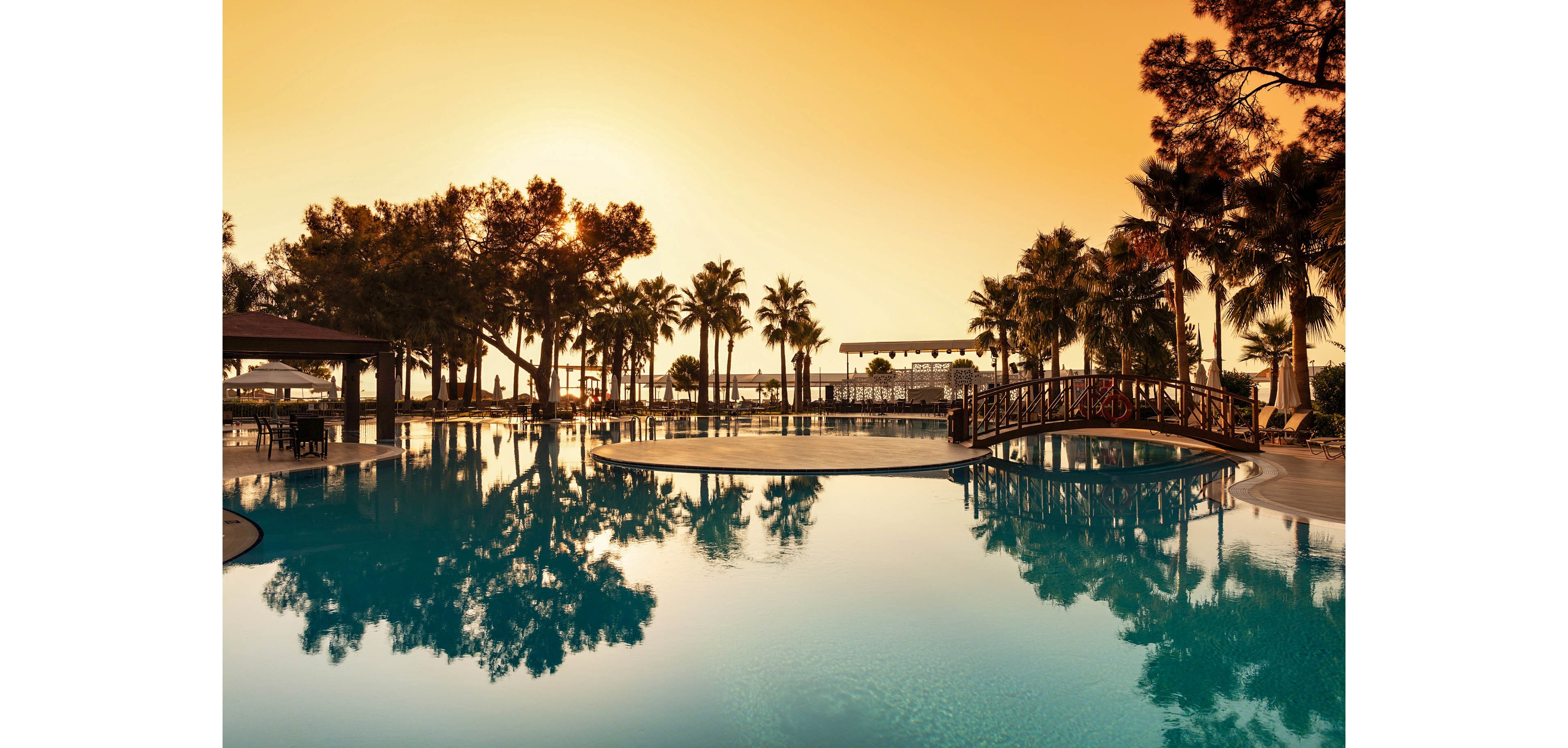 A large resort swimming pool reflects tall palm trees and an orange sunset sky. A small arched wooden bridge crosses part of the pool, adding a focal point to the tranquil scene. Lounge areas and structures are visible in the background, suggesting a vacation or holiday setting. The warm golden light and calm water create a peaceful, relaxing atmosphere.