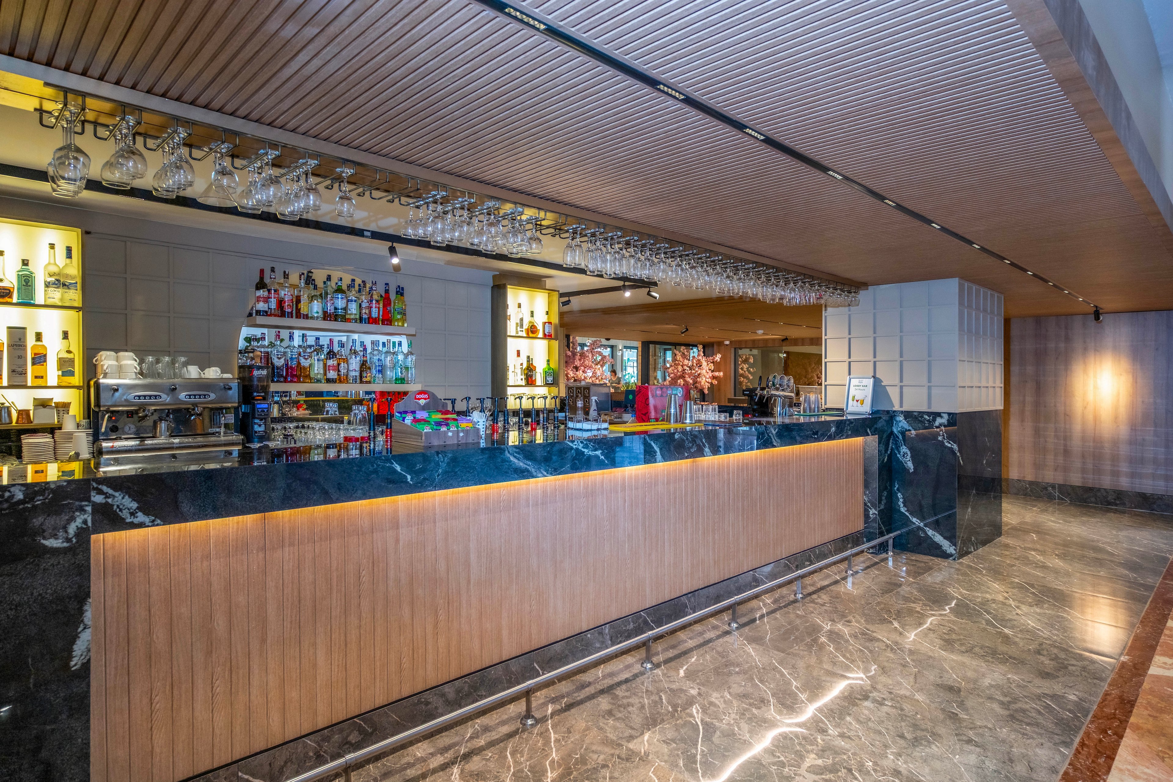 A contemporary bar interior features a long marble and wood counter with a sleek, minimalist design. Shelves behind the bar display an assortment of bottles and glassware, while hanging glasses line the ceiling above the counter. Warm recessed lighting highlights the polished stone floor and wooden ceiling panels. The space appears clean, organized, and ready for guests in a hotel or upscale lounge setting.