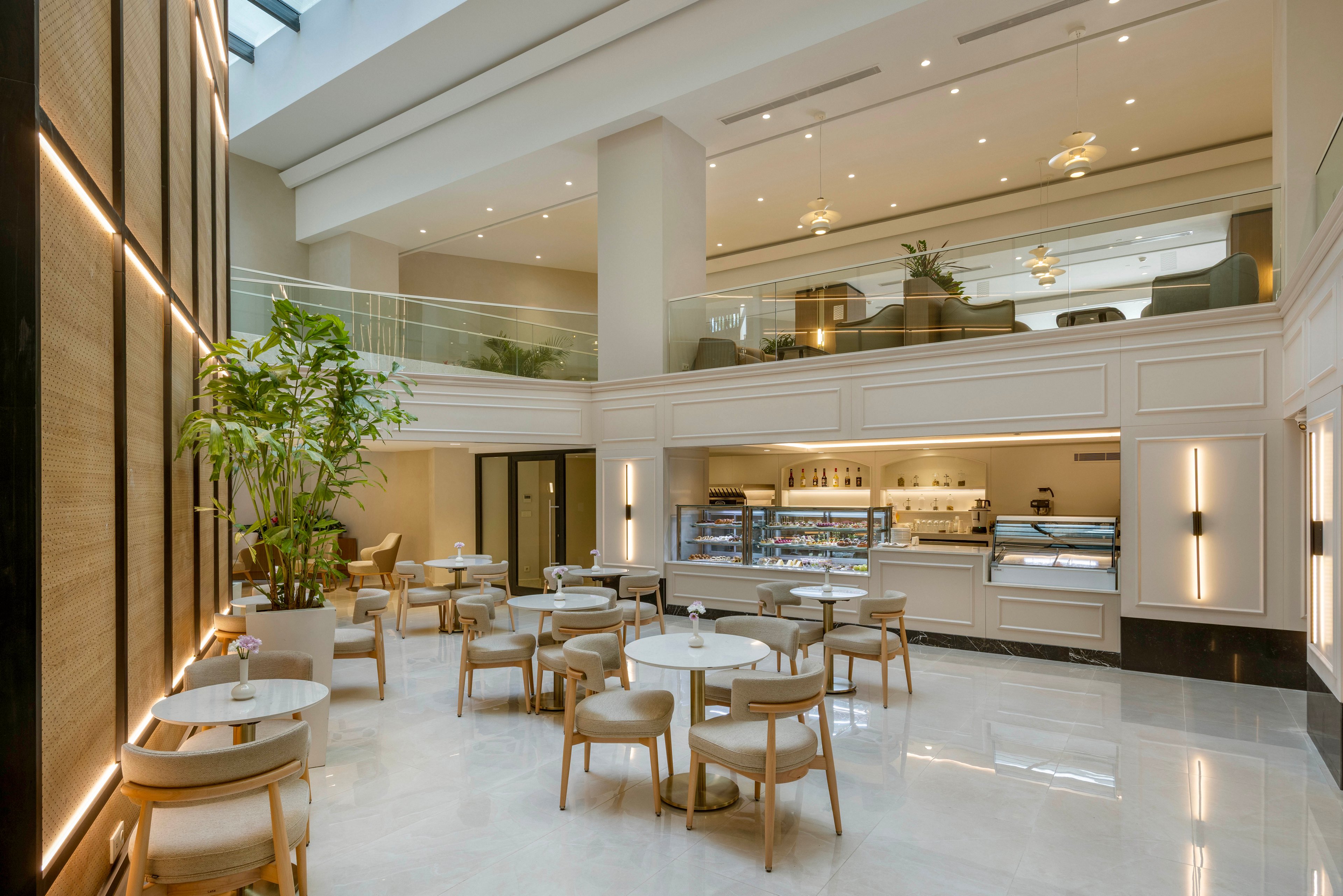 A spacious, modern café lounge features light-colored tables and chairs arranged neatly on a glossy floor. The setting includes a pastry and beverage counter with glass display cases and a barista station. Warm recessed lighting, tall columns, and a mezzanine level with additional seating create an airy, upscale atmosphere. Large indoor plants and natural light from the skylight enhance the calm, inviting mood.