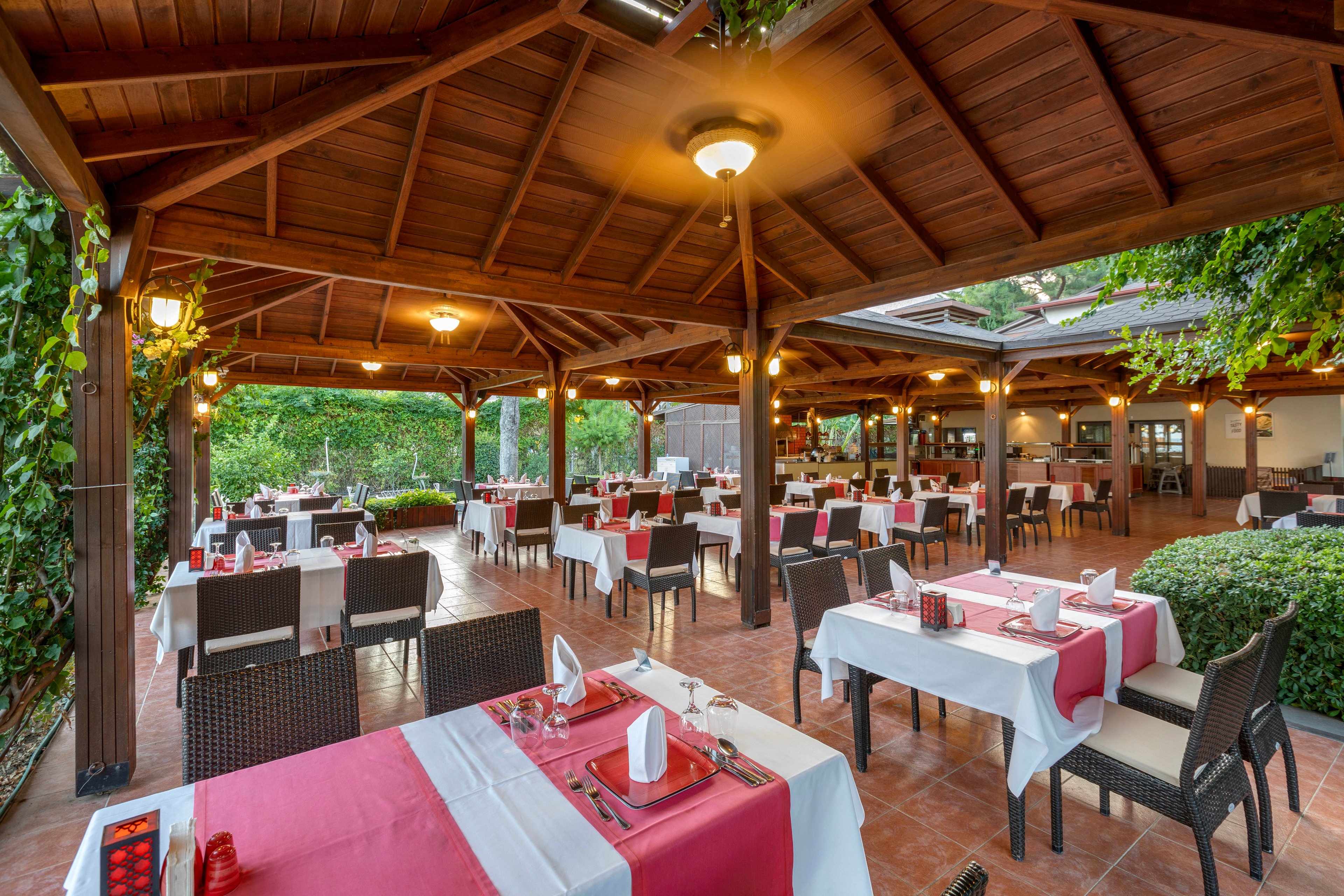 An open-air restaurant dining area is arranged under a large wooden pergola. Tables are neatly set with white and red tablecloths, napkins, and glassware, creating an elegant yet relaxed atmosphere. The space is surrounded by greenery and plants, adding a natural garden feel. Warm lighting and ceiling fans contribute to a comfortable, inviting setting for guests.
