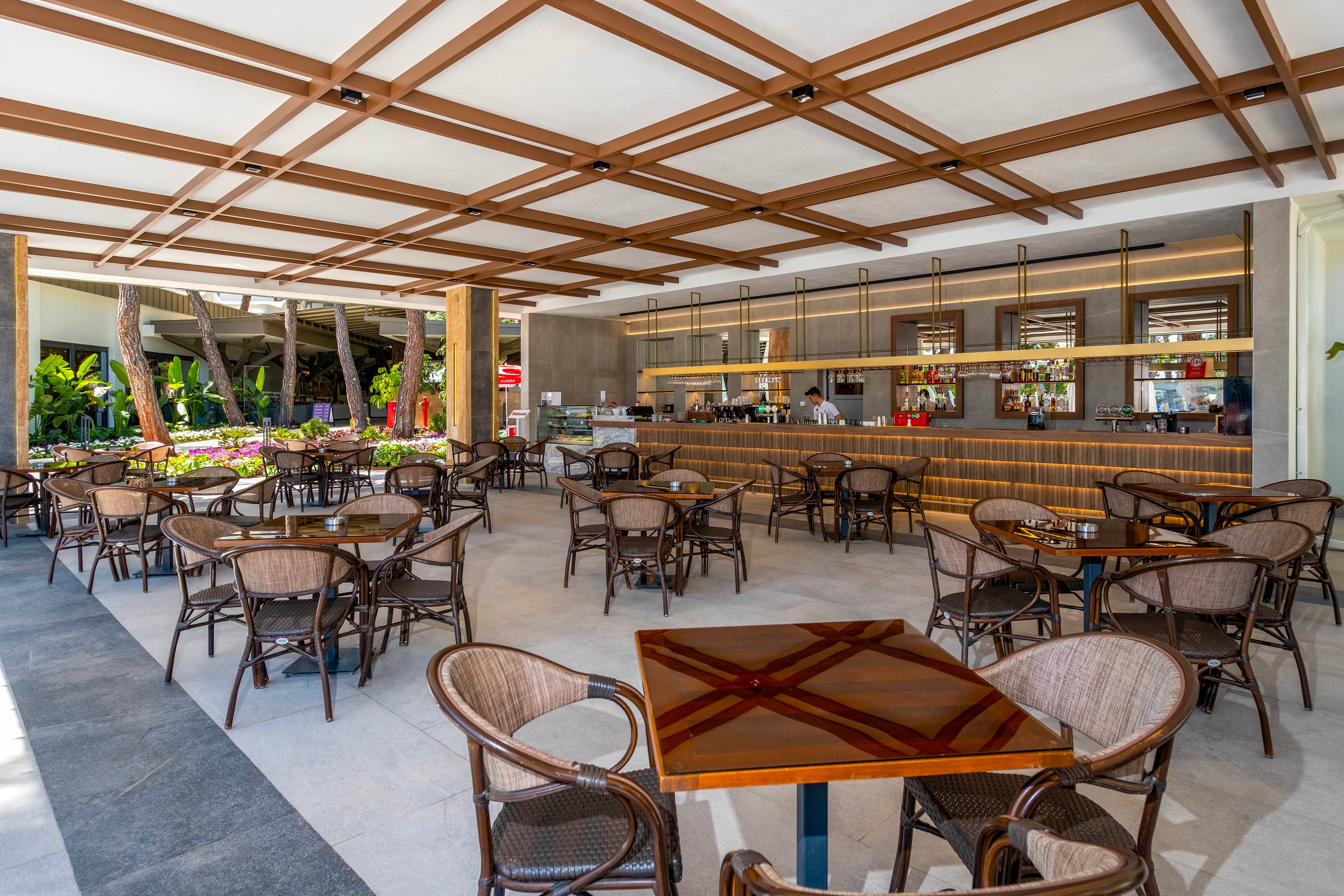 An open air bar area features numerous empty tables and wicker chairs neatly arranged. The bar counter stretches along the back wall with bottles, glasses, and equipment visible on the shelves. Wooden beams create a geometric pattern on the ceiling, adding structure and warmth to the space. Tropical greenery and bright daylight outside suggest a relaxed resort or hotel setting.