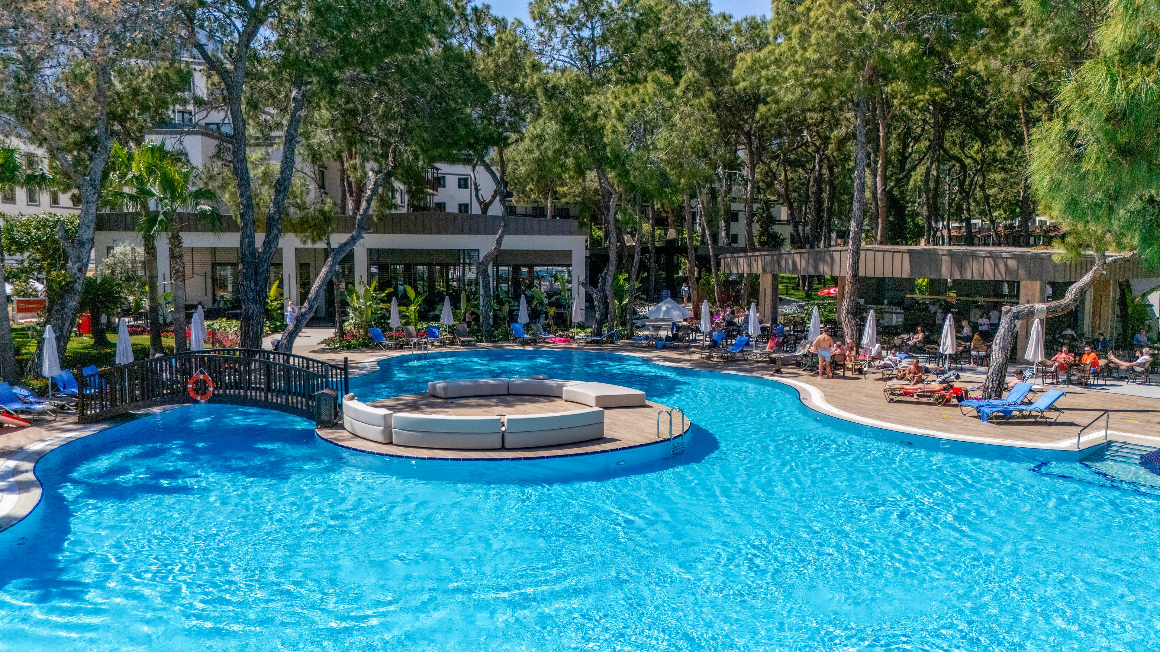 Large curved outdoor swimming pool at a holiday resort is shown under bright sunshine. Sun loungers and umbrellas line the poolside where guests relax and enjoy the warm weather. A circular seating island sits in the center of the pool, adding a focal point to the scene. Tall green trees and low-rise resort buildings create a tranquil, shaded backdrop.