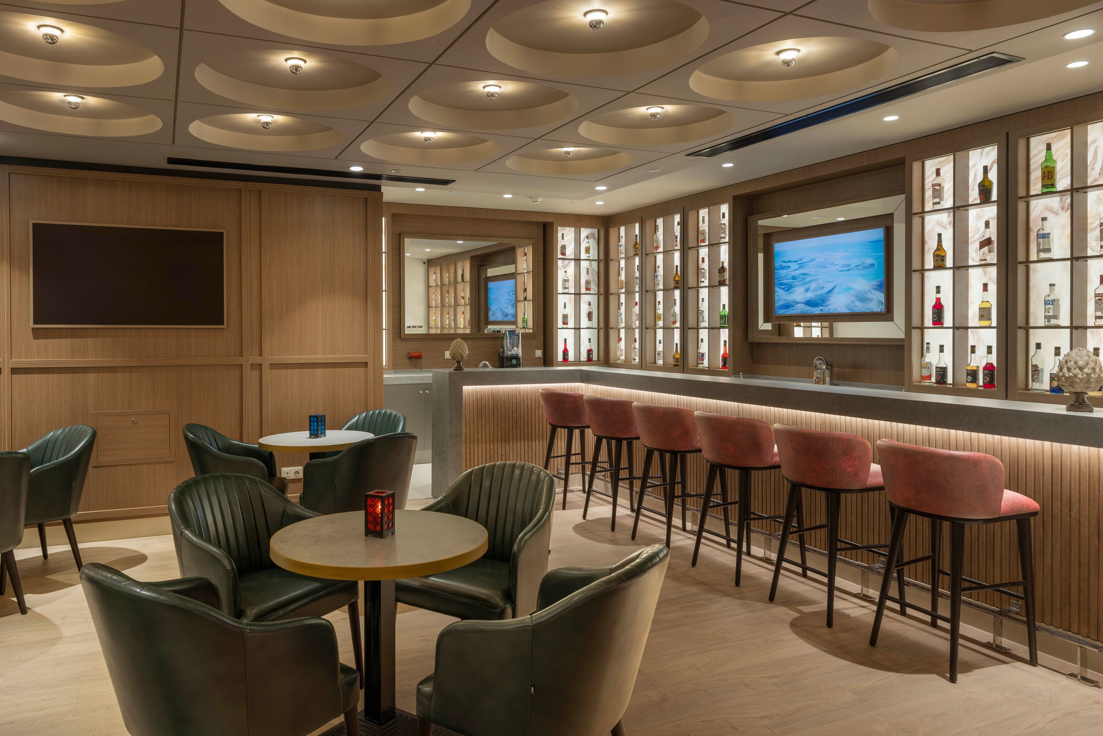 A contemporary lounge bar features a sleek counter with high stools and a wall of illuminated shelves displaying assorted bottles. In the foreground, round tables are surrounded by upholstered armchairs, creating an intimate seating area. Warm wood paneling, recessed ceiling lights, and soft accent lighting contribute to a cozy yet upscale atmosphere. A large flat-screen television and decorative candle holders add to the modern hospitality setting.