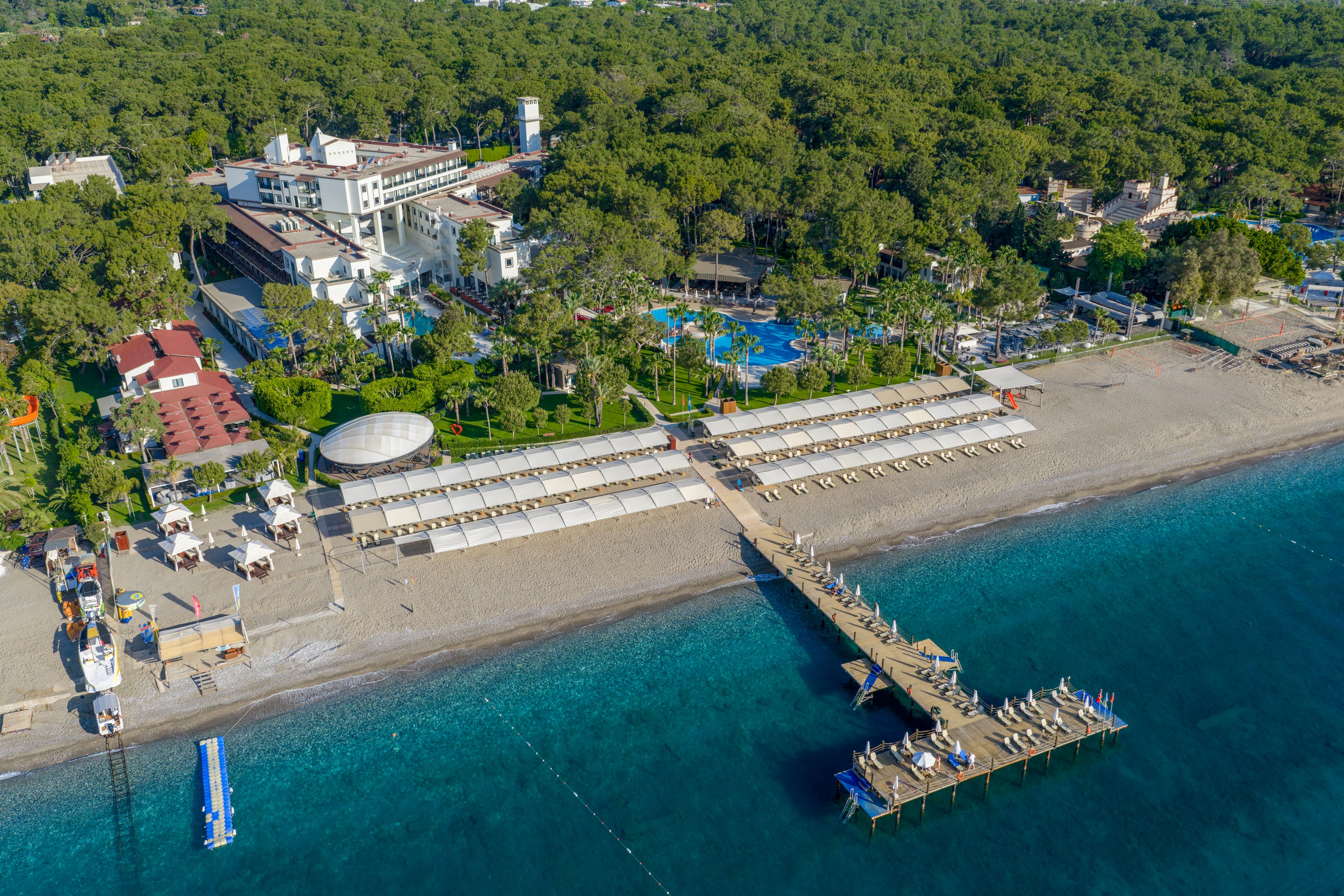 A coastal resort complex is seen from above, nestled among dense green trees beside a long sandy beach. A wooden pier extends out into clear turquoise water, with sun loungers and umbrellas arranged along the shore. Several buildings, pools, and recreational areas are visible within the resort grounds. The scene suggests a tranquil vacation destination with organized beachfront facilities and calm sea conditions.