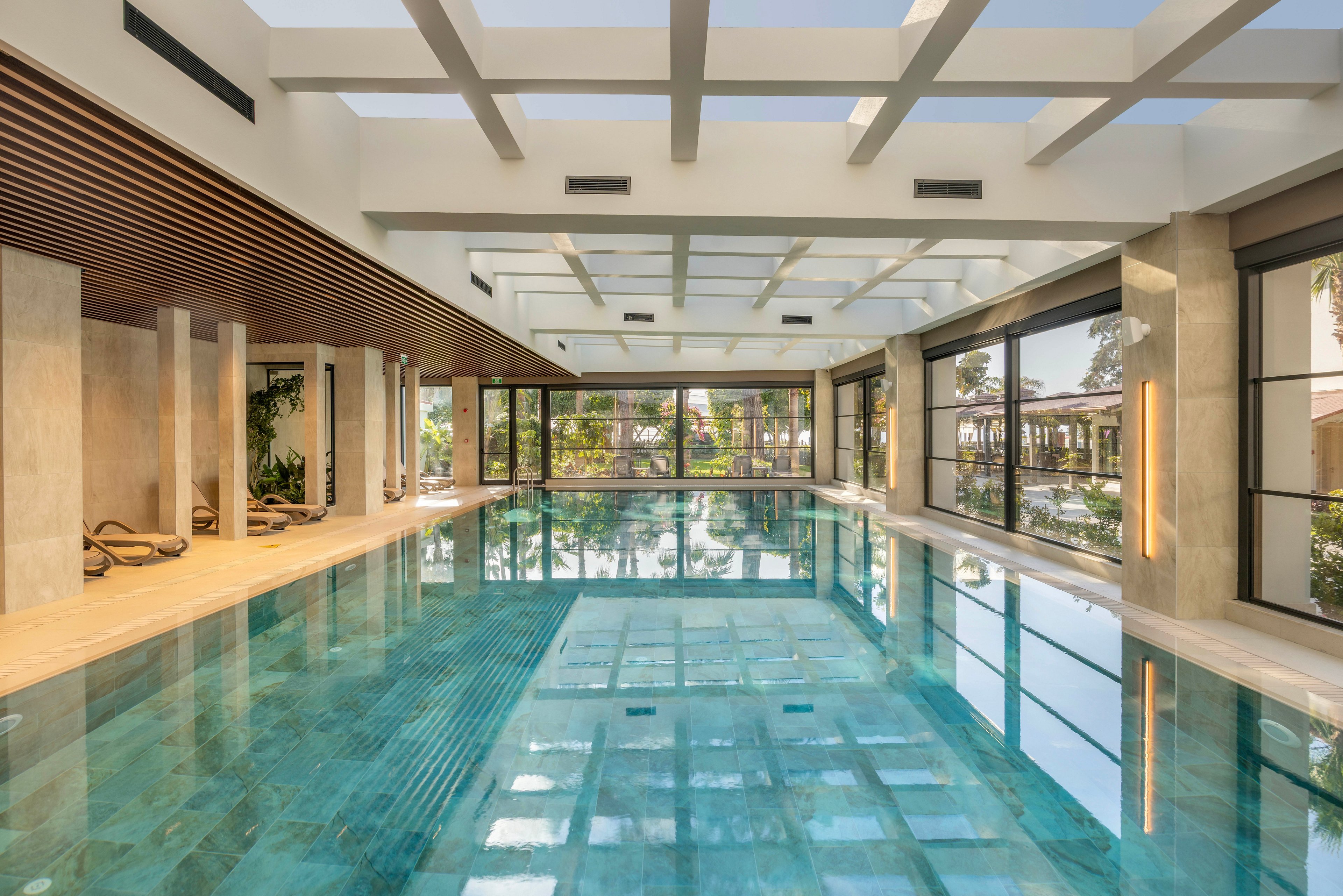 A spacious indoor swimming pool is featured in a contemporary building with clean architectural lines. Large floor-to-ceiling windows on both sides reveal greenery and outdoor structures, filling the space with natural light. The clear blue water reflects the ceiling grid and surrounding columns, creating a calm and inviting atmosphere. Lounge chairs and subtle lighting along the walls enhance the relaxing, resort-like setting.