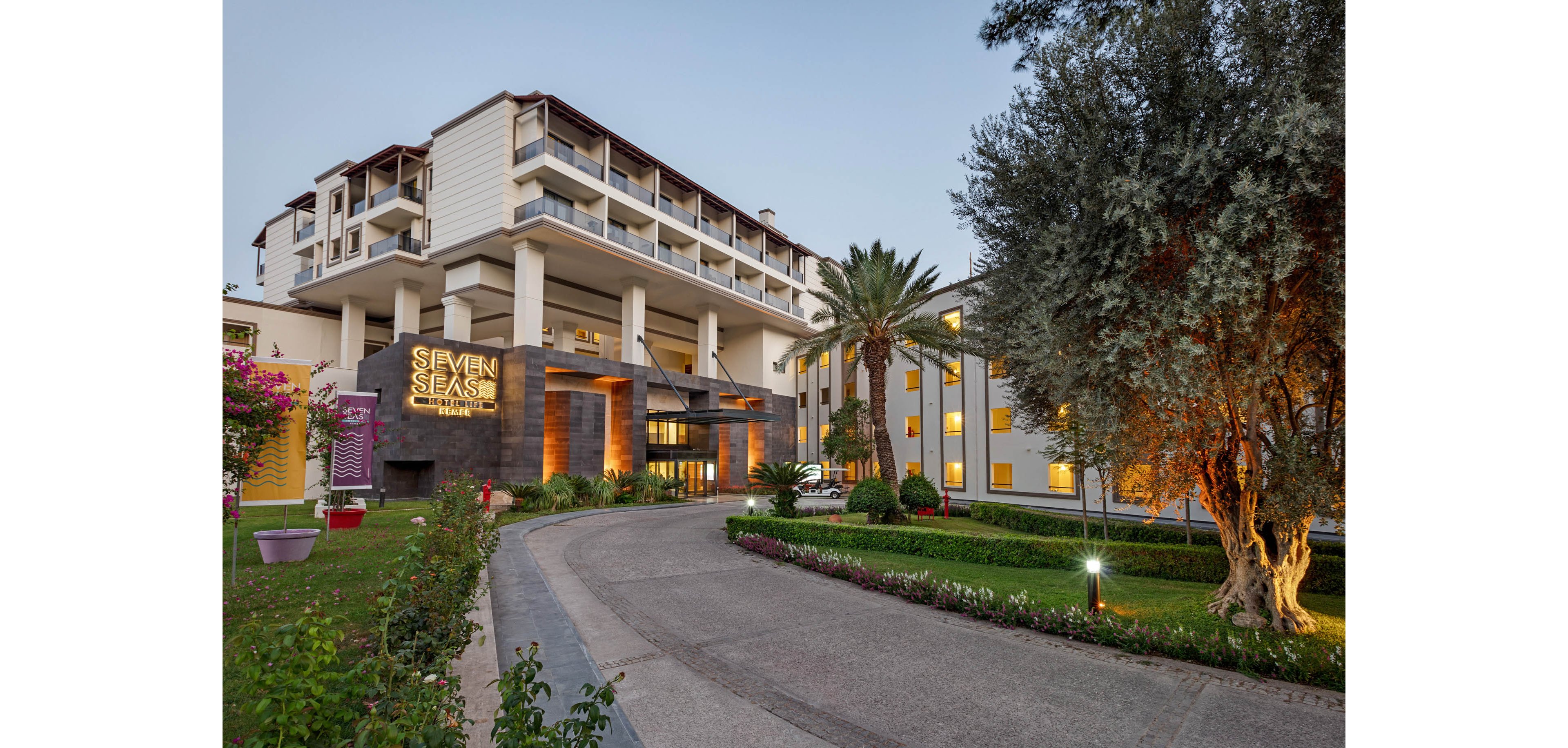 A contemporary multi-story resort hotel is shown from the driveway at dusk. Warm lights glow from the entrance and guestroom windows, creating an inviting atmosphere. The landscaped grounds feature green lawns, flowering plants, and palm trees along the curved drive. A large illuminated sign reading "SEVEN SEAS" is prominently displayed near the main entrance.