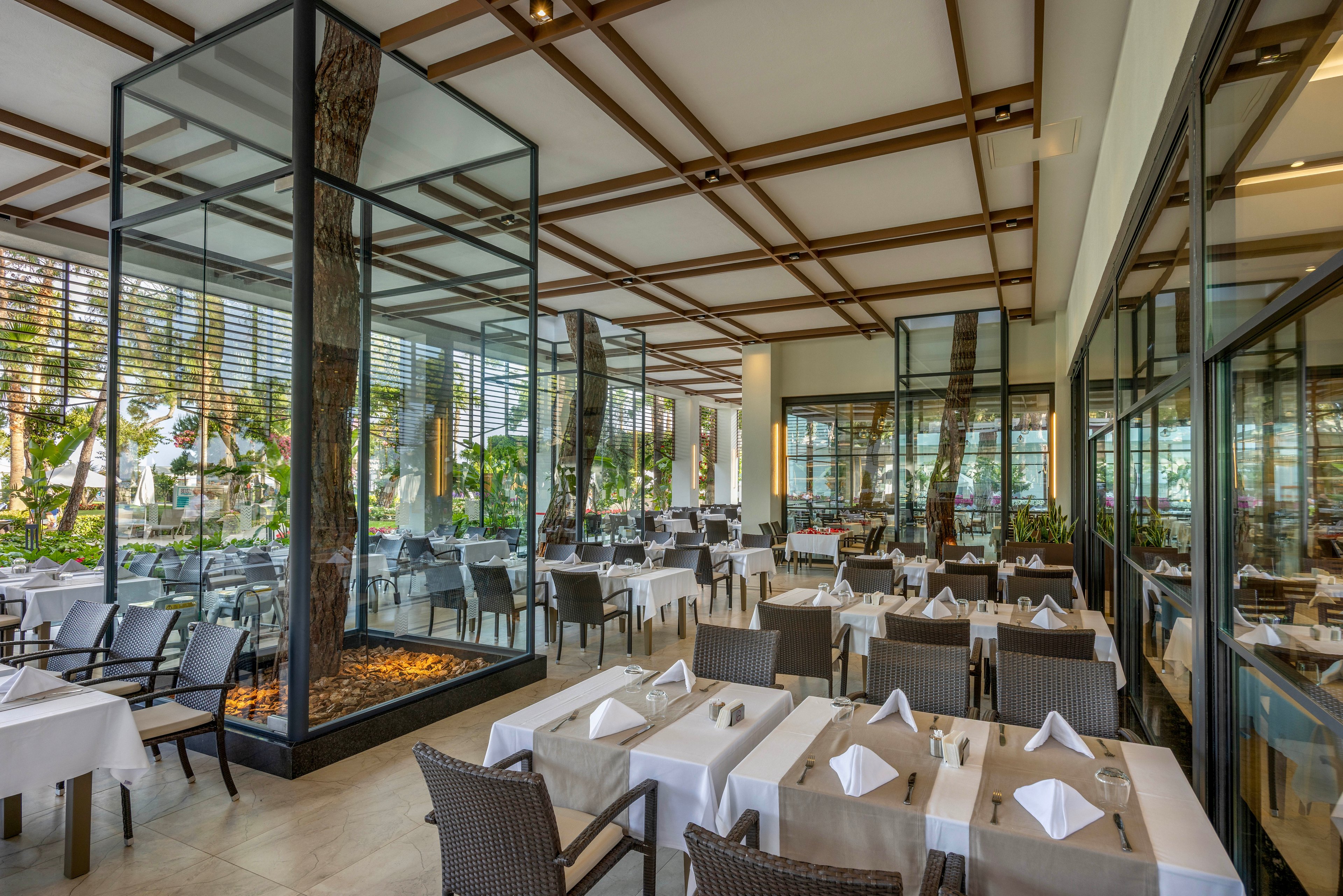 A contemporary restaurant dining area features neatly arranged tables covered with white tablecloths and folded napkins. Wicker-style chairs surround the tables, creating a comfortable yet upscale atmosphere. Large glass enclosures with trees and decorative stones act as striking design elements in the center of the space. Floor-to-ceiling windows and open sides reveal lush greenery outside, giving the restaurant an airy, resort-like feel.