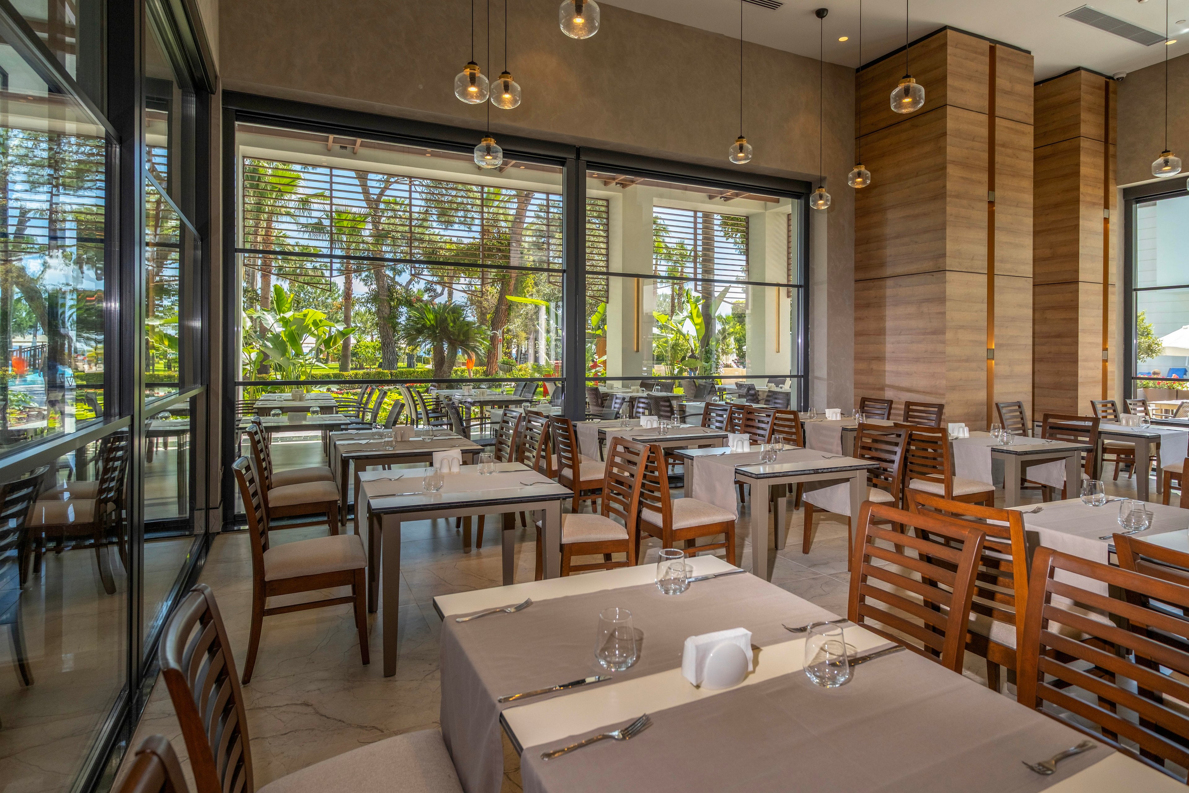 A contemporary restaurant dining area features neatly arranged wooden chairs and tables covered with beige tablecloths. Large floor-to-ceiling windows open to a lush tropical garden and outdoor seating area. Pendant lights hang from the high ceiling, adding a warm glow to the neutral-toned interior. Table settings with glassware and napkins suggest a prepared yet unoccupied dining space.