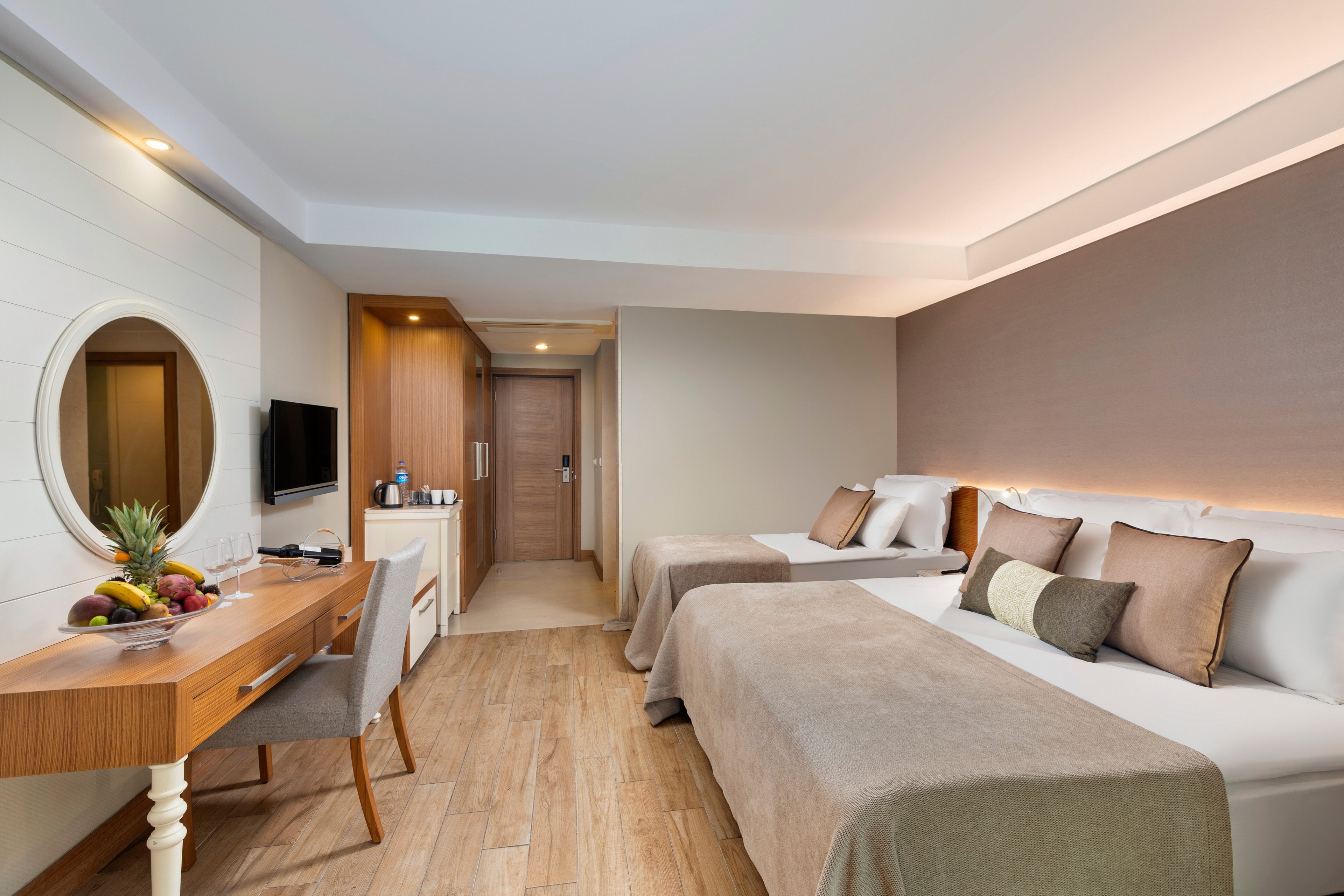 A contemporary hotel room featuring two neatly made twin beds with beige bedding and accent pillows. The setting includes a wooden desk with a mirror, a television, and a fruit arrangement. The room has warm lighting and a minimalist design, with wooden flooring and neutral tones creating a cozy atmosphere.