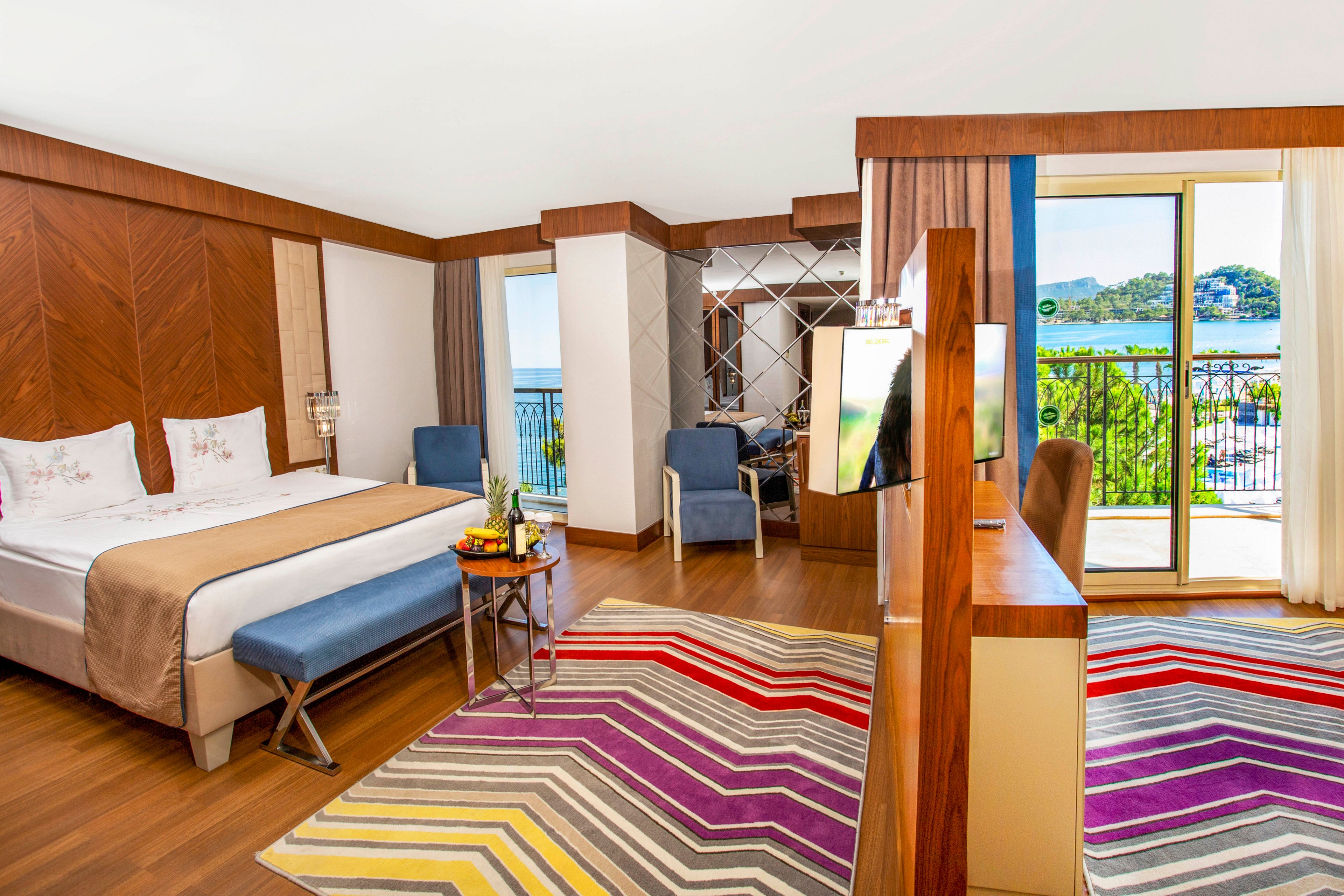 A contemporary hotel room features a large bed with crisp white linens and neutral bedding. The space includes a sitting area with blue armchairs and a television divider separating the sleeping and lounge zones. A vibrant multicolored striped rug adds energy to the wooden floor, while large windows and a balcony door open to a scenic outdoor view. Natural light brightens the room, highlighting the warm wood finishes and modern decor.