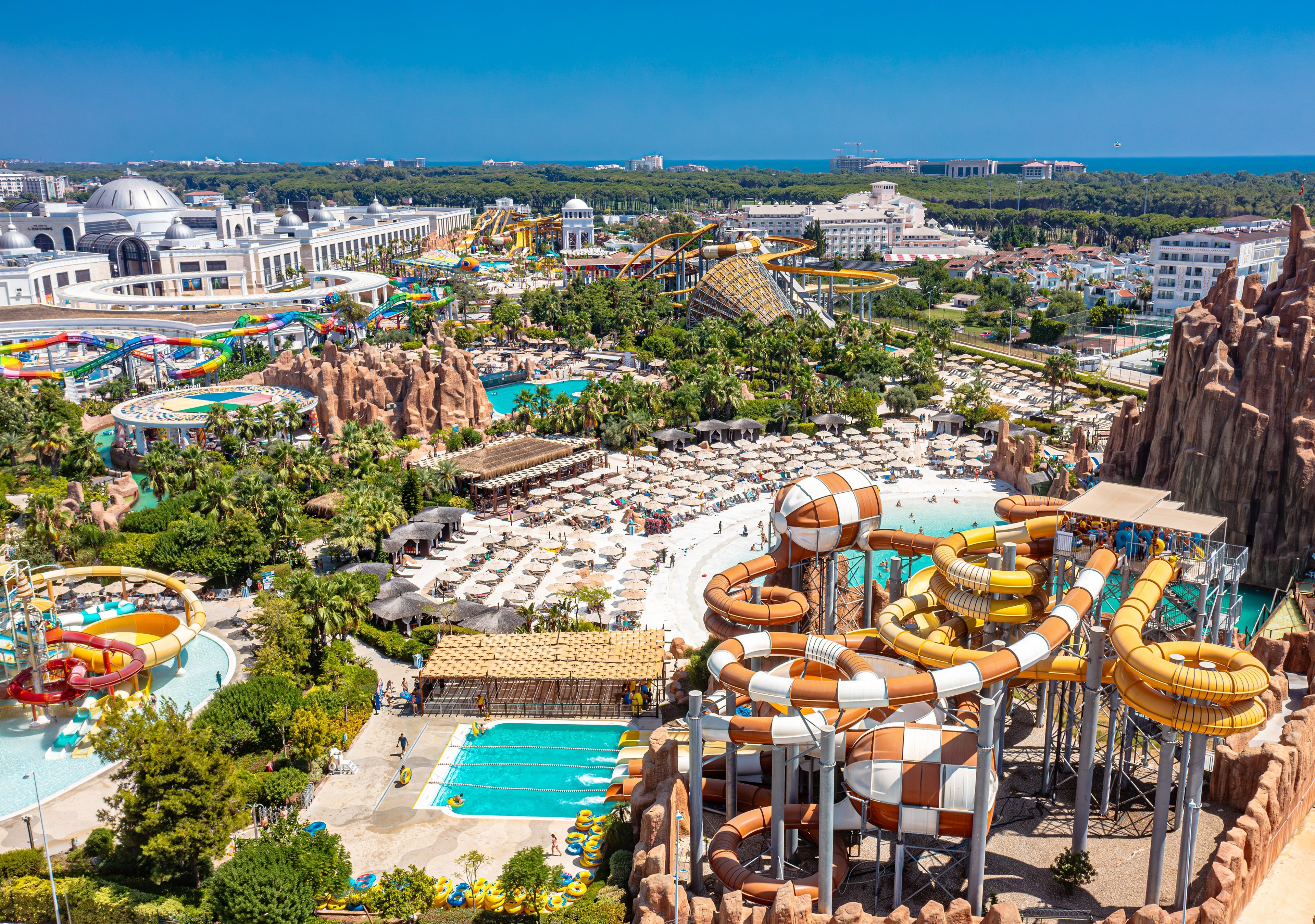 Large outdoor water park with numerous slides and pools is seen from an elevated viewpoint. The complex features twisting tube slides, funnel slides, and lazy rivers surrounded by lush greenery. Rows of sun loungers and umbrellas fill the central area, indicating a busy leisure destination. In the distance, the coastline and blue sea frame the expansive resort setting under a clear sky.