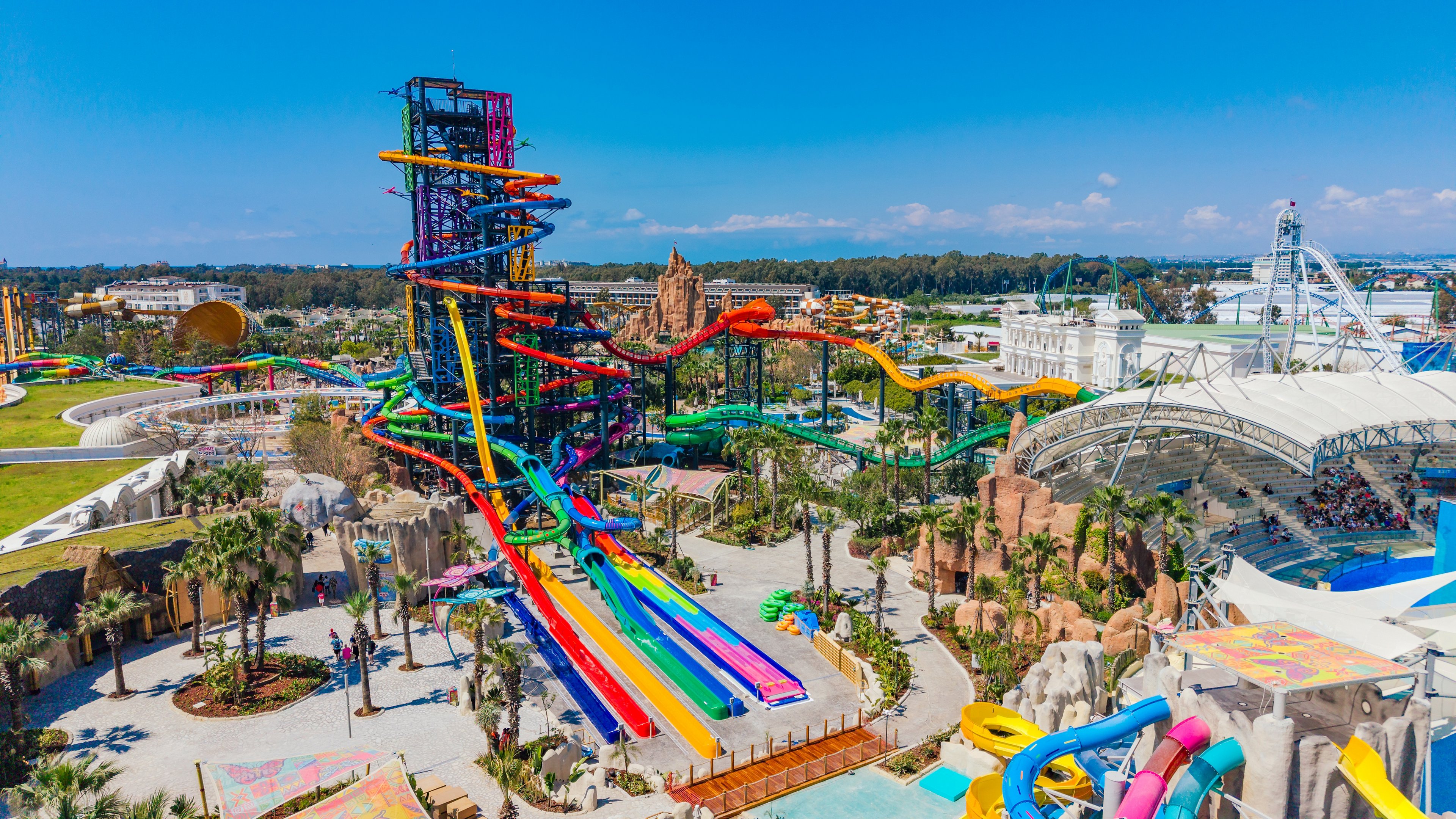 A large water park features a towering structure of multicolored water slides dominating the scene. Numerous winding tubes and straight slides descend into splash pools surrounded by palm trees and landscaped walkways. Visitors can be seen enjoying the attractions under a bright blue sky with scattered clouds. The overall atmosphere is vibrant, sunny, and energetic, suggesting a fun summer destination.