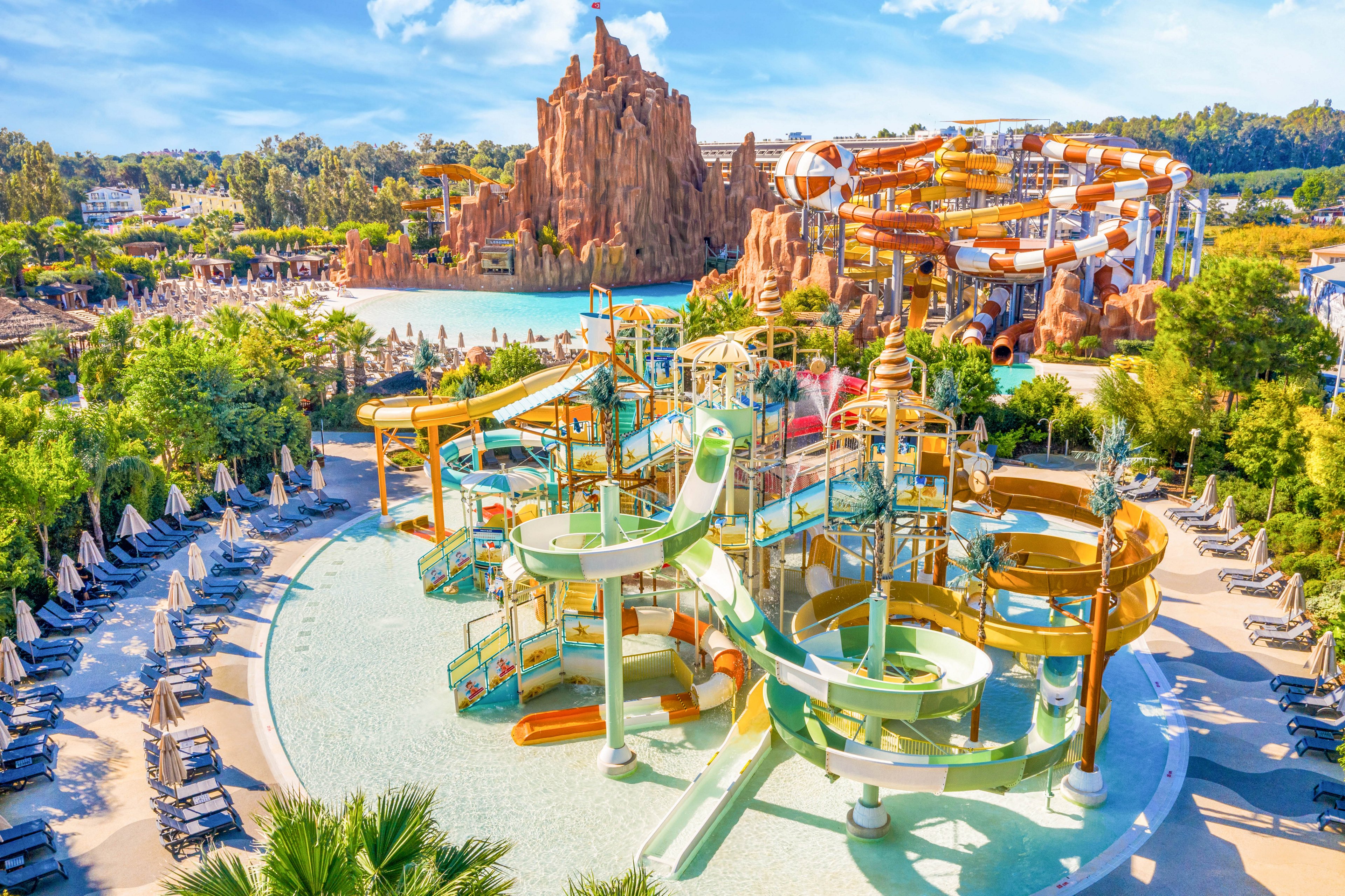 A large outdoor water park features multiple colorful water slides and play structures in a shallow pool. Surrounding the central attraction are rows of lounge chairs and umbrellas arranged neatly for visitors. In the background, additional tall slides and a rocky themed structure rise above the trees. The scene is bright and sunny, emphasizing a fun, family-friendly vacation atmosphere.