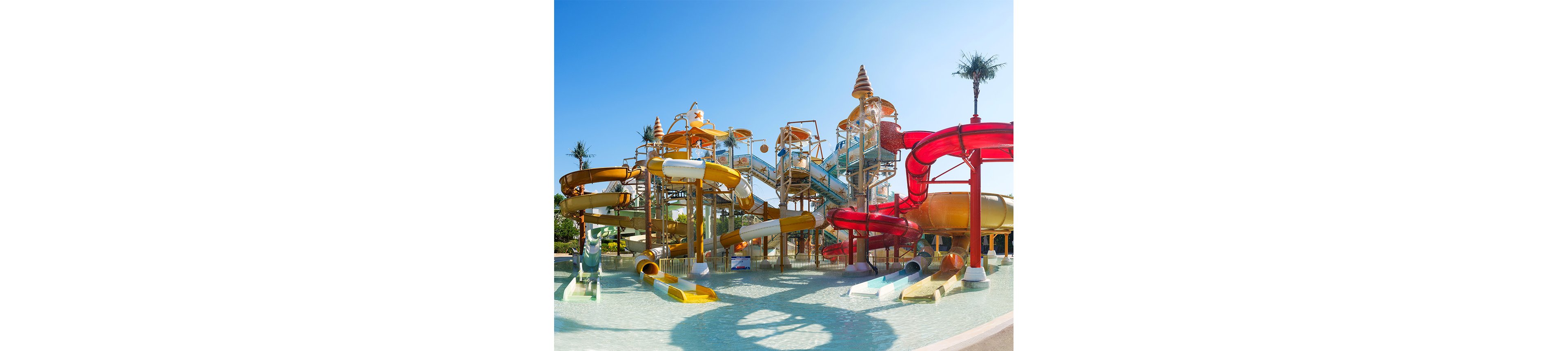 A large water park structure features multiple colorful twisting slides emptying into a shallow pool. Elevated walkways and staircases connect the slides, with water jets and splash elements visible throughout. The scene is set outdoors under a clear blue sky with a few palm trees in the background. Bright colors and sparkling water create a fun, playful atmosphere suitable for families and children.