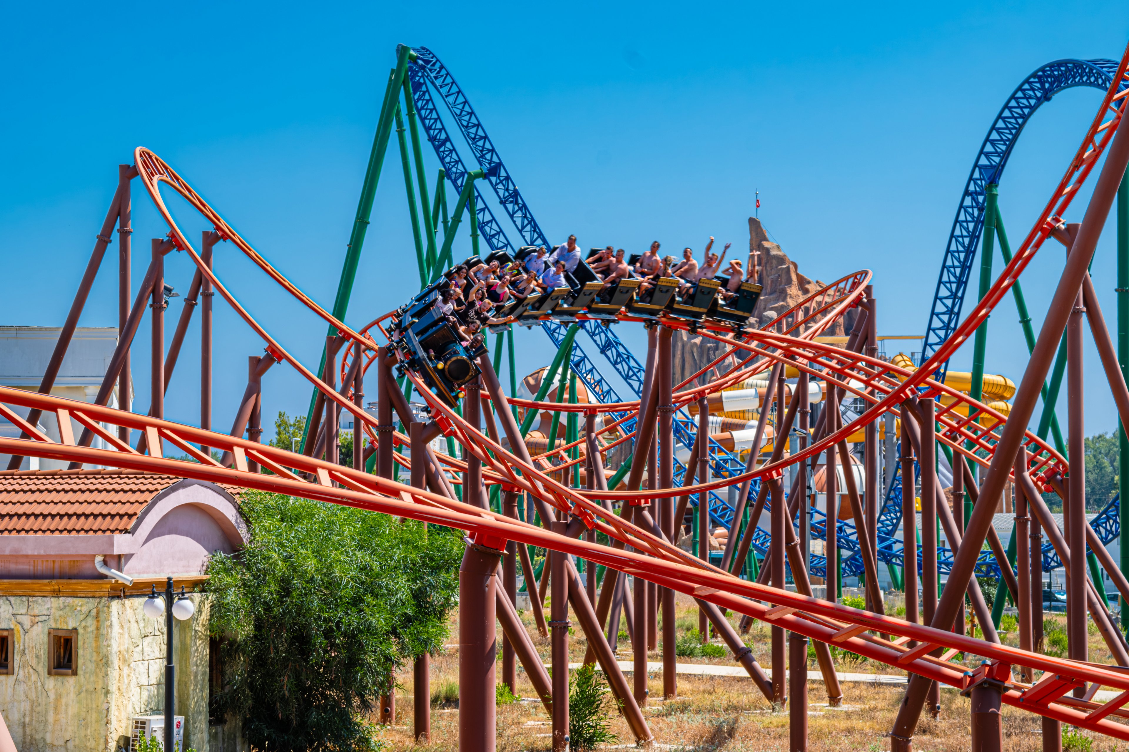 A train full of riders speeds along a twisting roller coaster track. The scene is set in an amusement park with multiple colorful steel tracks crossing each other. People raise their hands as the coaster leans into a curve, emphasizing motion and excitement. Bright blue sky and warm sunlight create a vibrant, summery atmosphere.