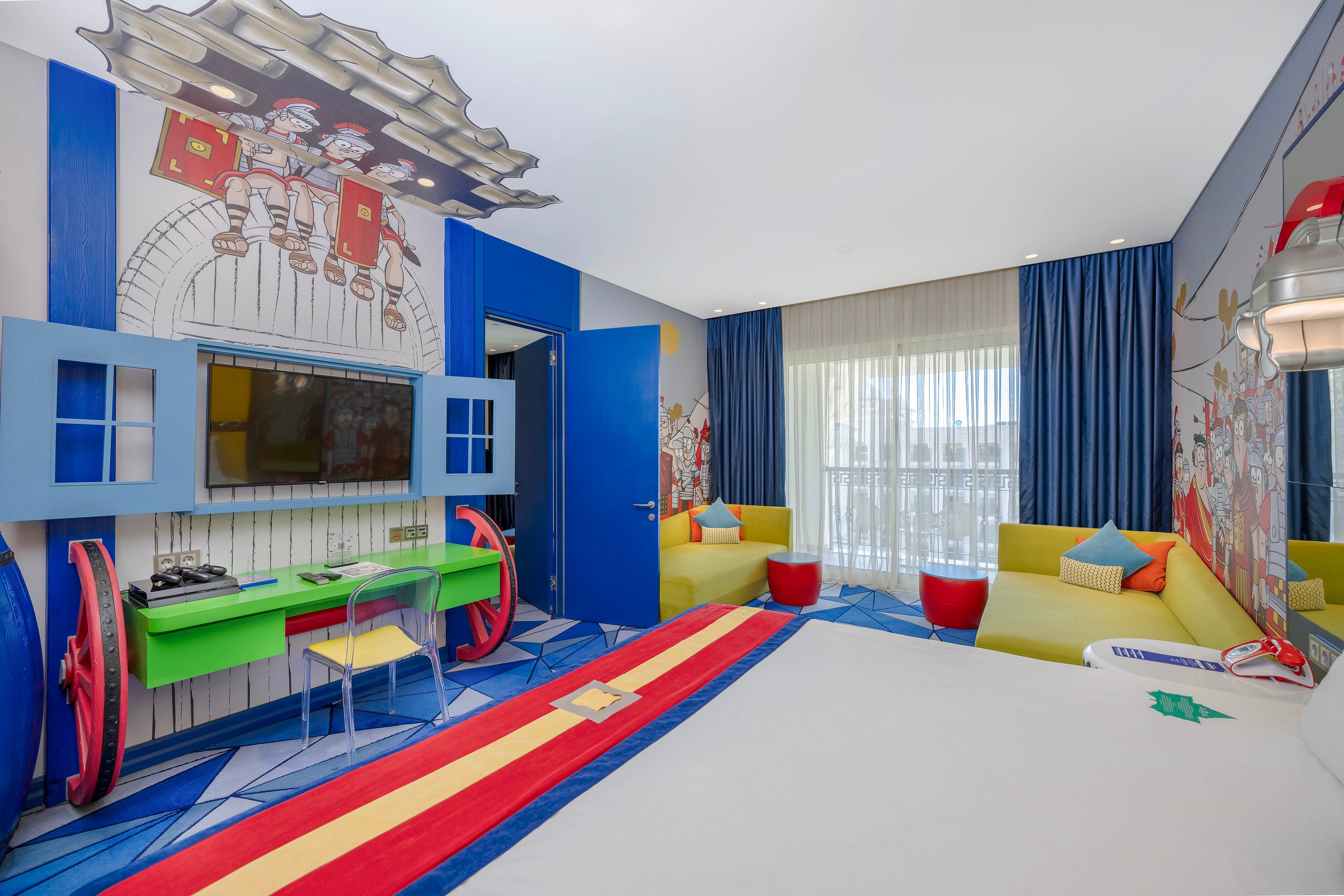 A vibrant hotel room features cartoon-inspired wall art and playful furniture. The setting includes a bed with a striped runner, a green desk with a transparent chair, and yellow sofas with red ottomans. Large windows with sheer curtains allow natural light to fill the space, while bold blue and red accents create a lively atmosphere. The room is designed to appeal to families and children, with whimsical visuals and a cheerful mood.